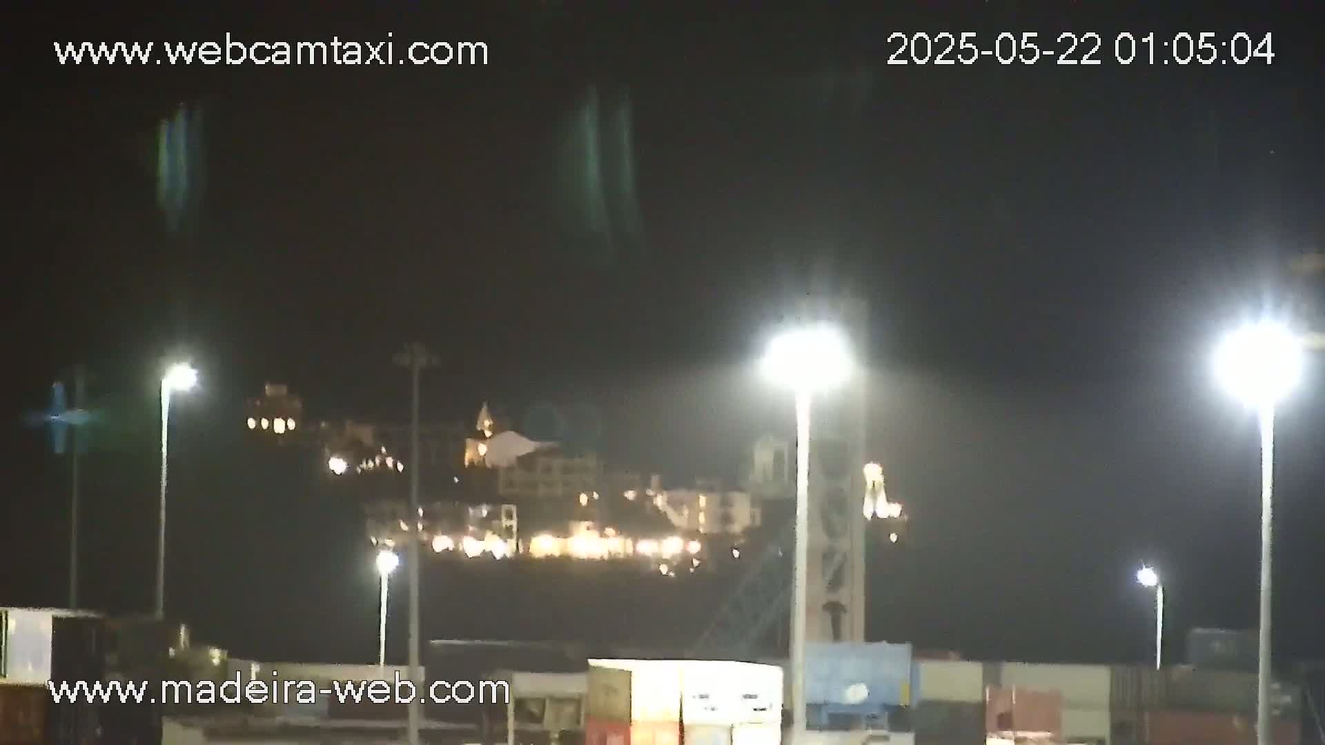 At night, a hilltop with illuminated buildings is visible beyond a brightly lit area containing cargo containers and tall light posts.