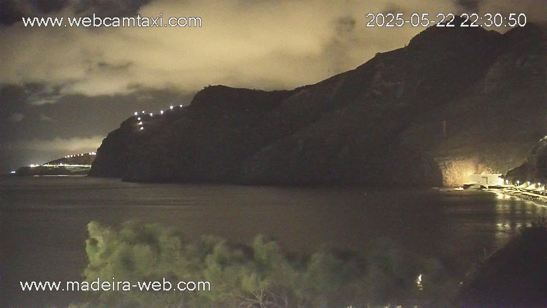 A nighttime coastal scene shows a dark mountain with lights along its edge, overlooking a calm body of water under a cloudy sky.