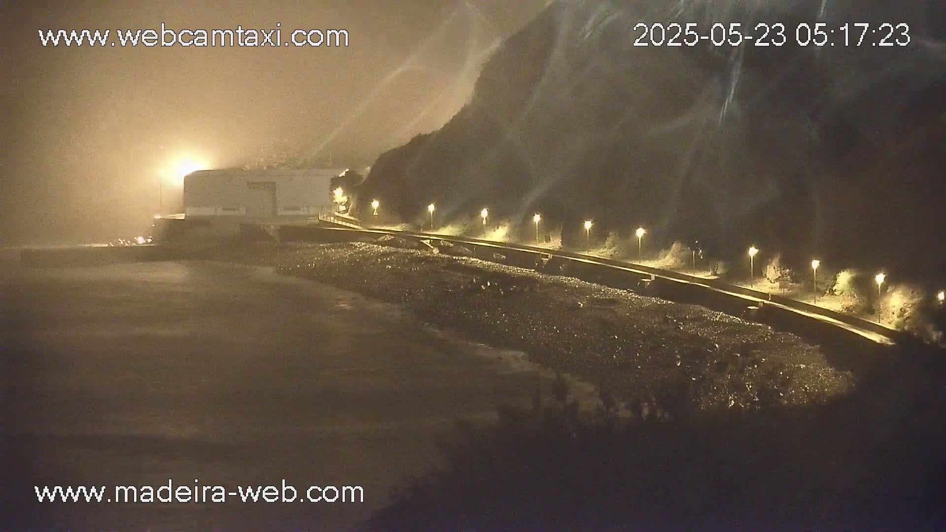 A nighttime coastal scene shows a building near a rocky shore with a roadway lined with lights, under hazy, windy conditions.