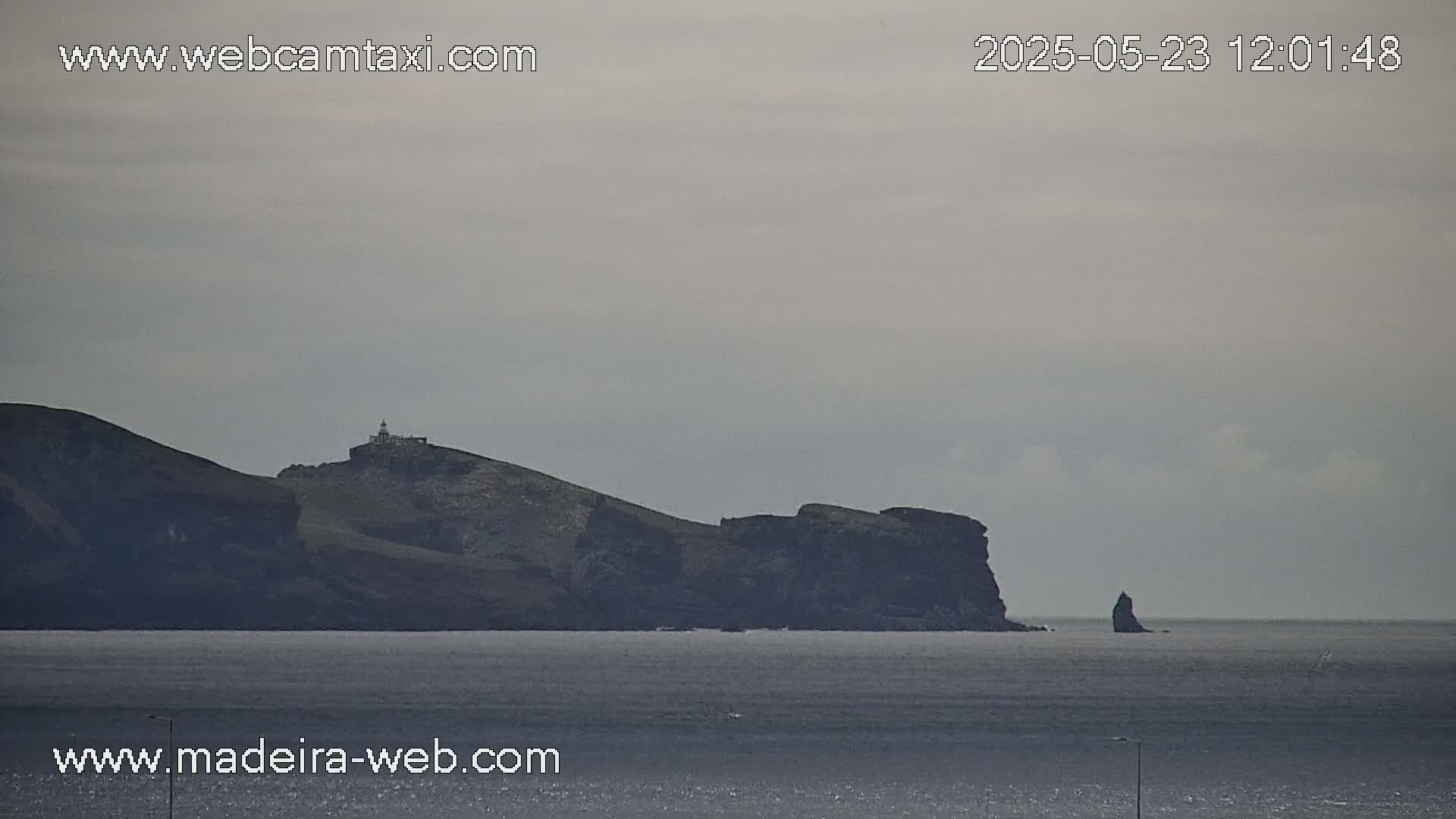A distant, dark island with a lighthouse sits on the horizon of a calm, gray ocean under an overcast sky.