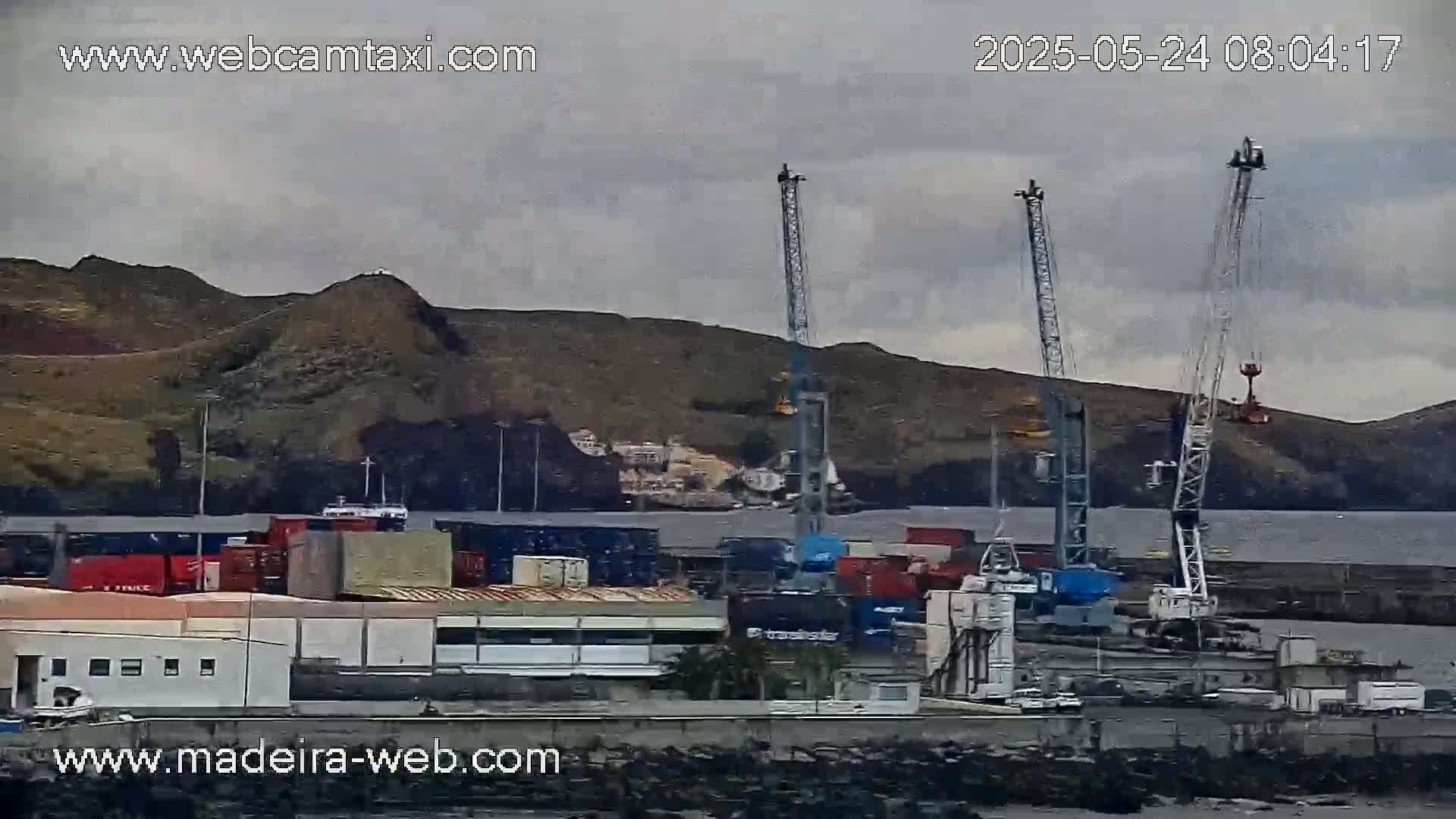 A cargo port with several cranes is shown under an overcast sky, with a dark hill in the background.