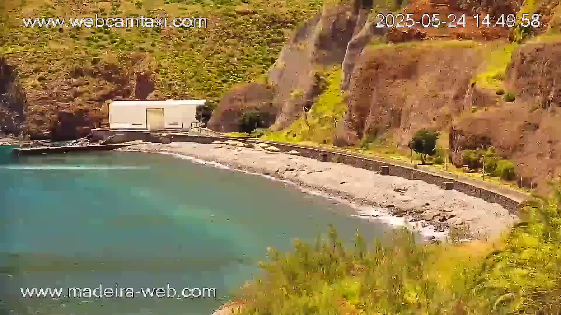 A rocky, pebble beach curves around a calm, teal bay next to a steep, brown cliff under a sunny sky.