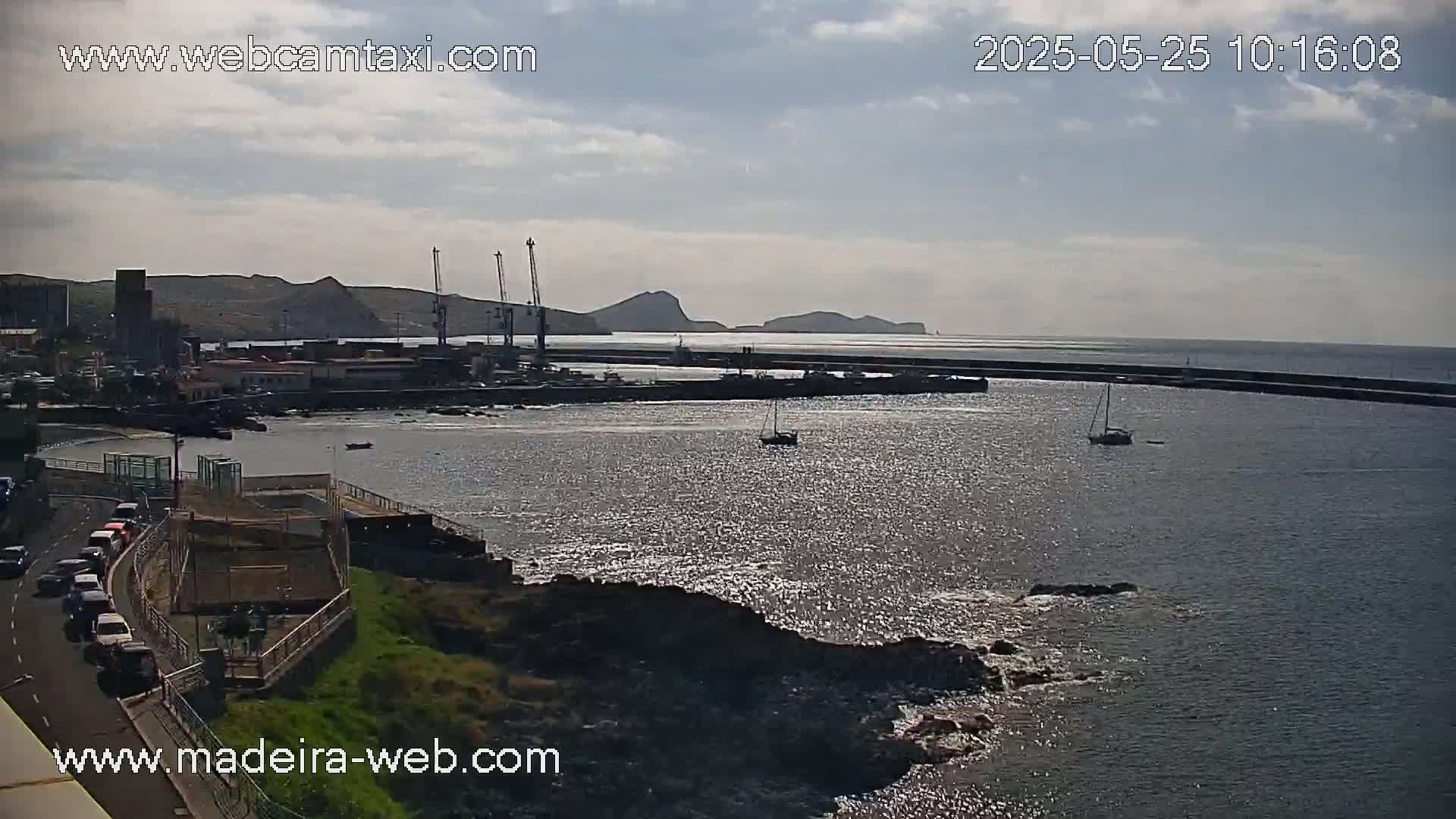 A harbor with several boats, a breakwater, and industrial buildings is visible under a partly cloudy sky.