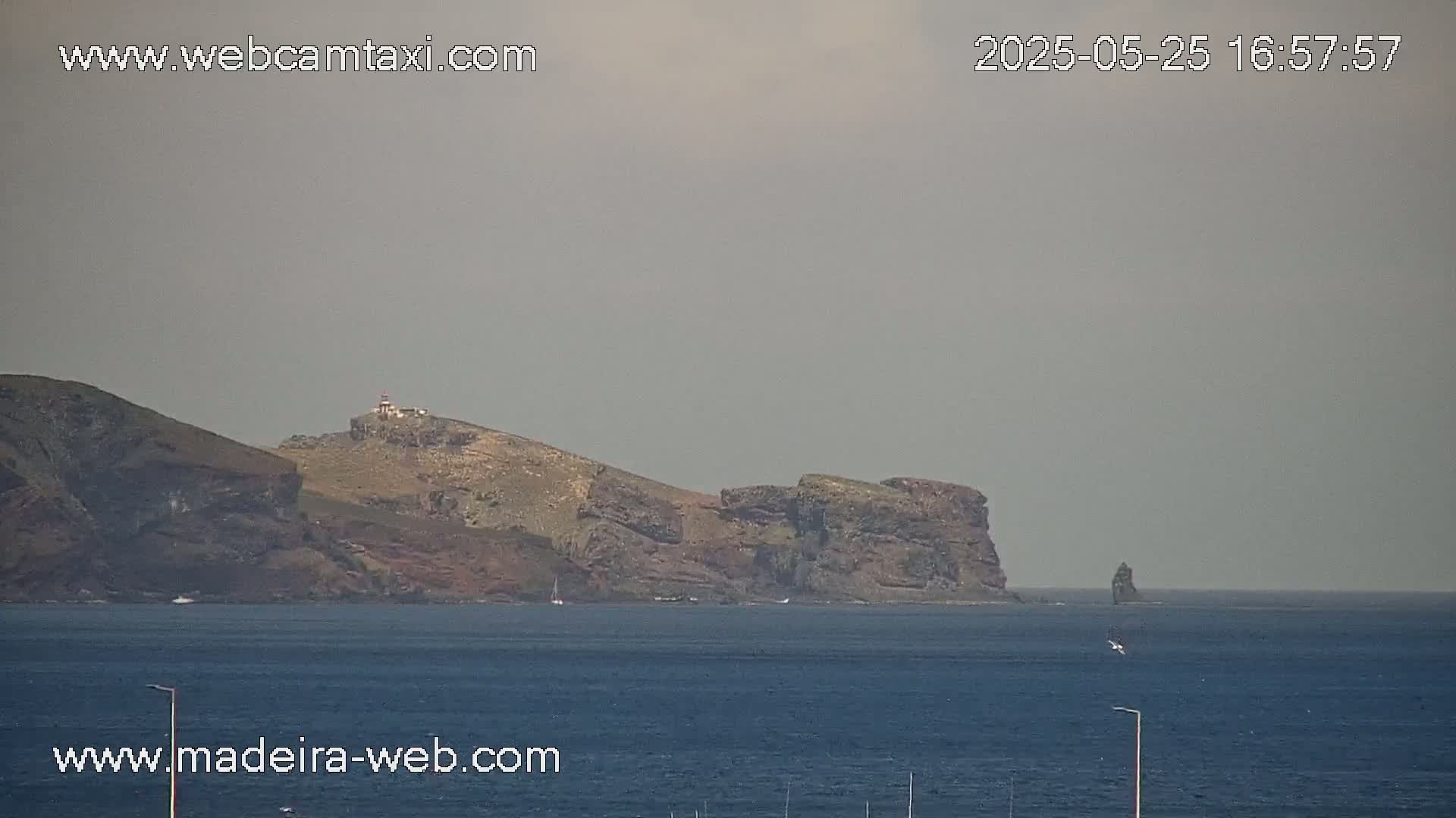 A hazy, long shot shows a rocky, mountainous coastline with a lighthouse, extending into a calm blue ocean with a solitary, dark rock formation in the distance.