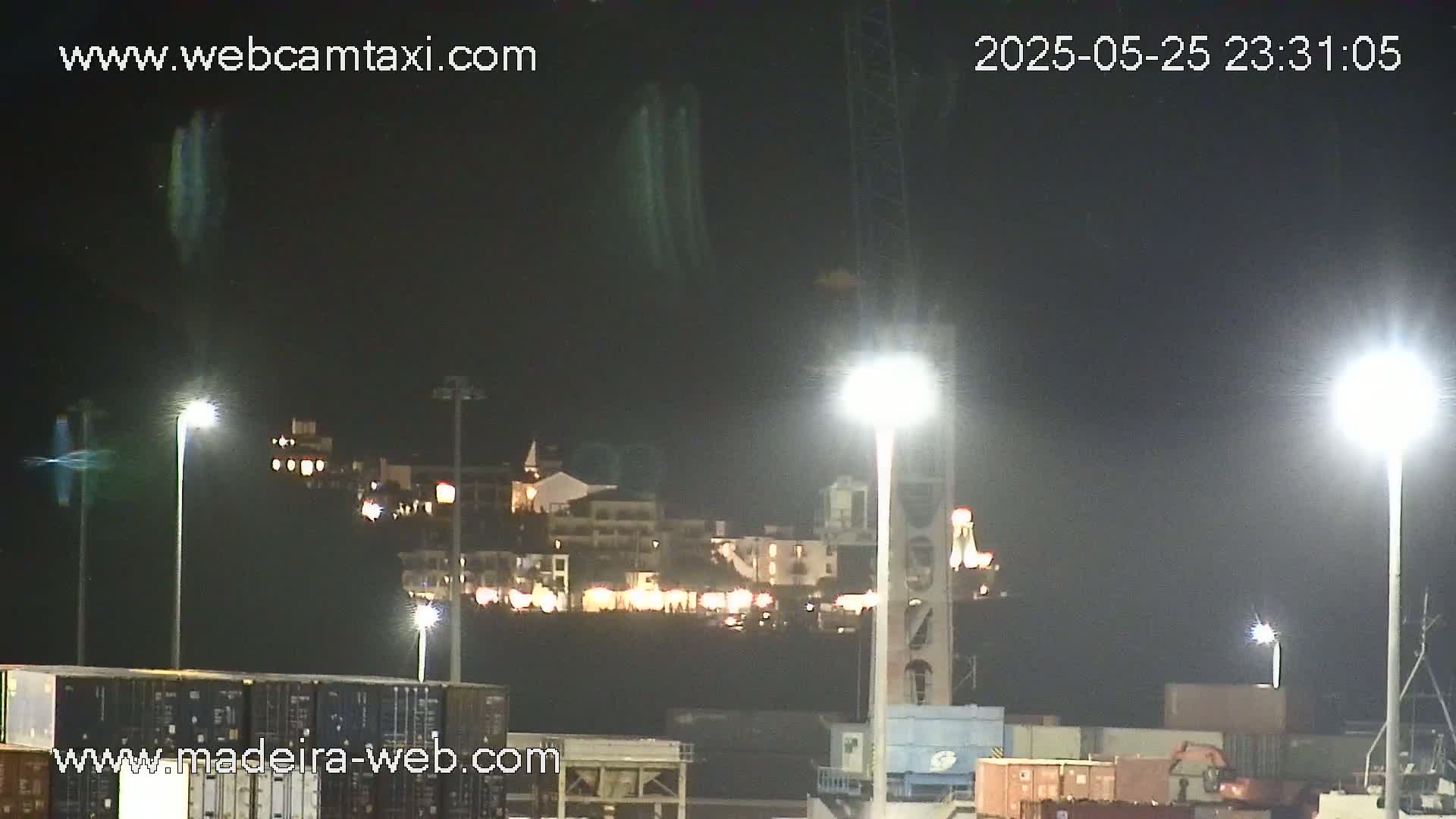 At night, a brightly lit waterfront area with buildings and shipping containers is visible under a dark sky.