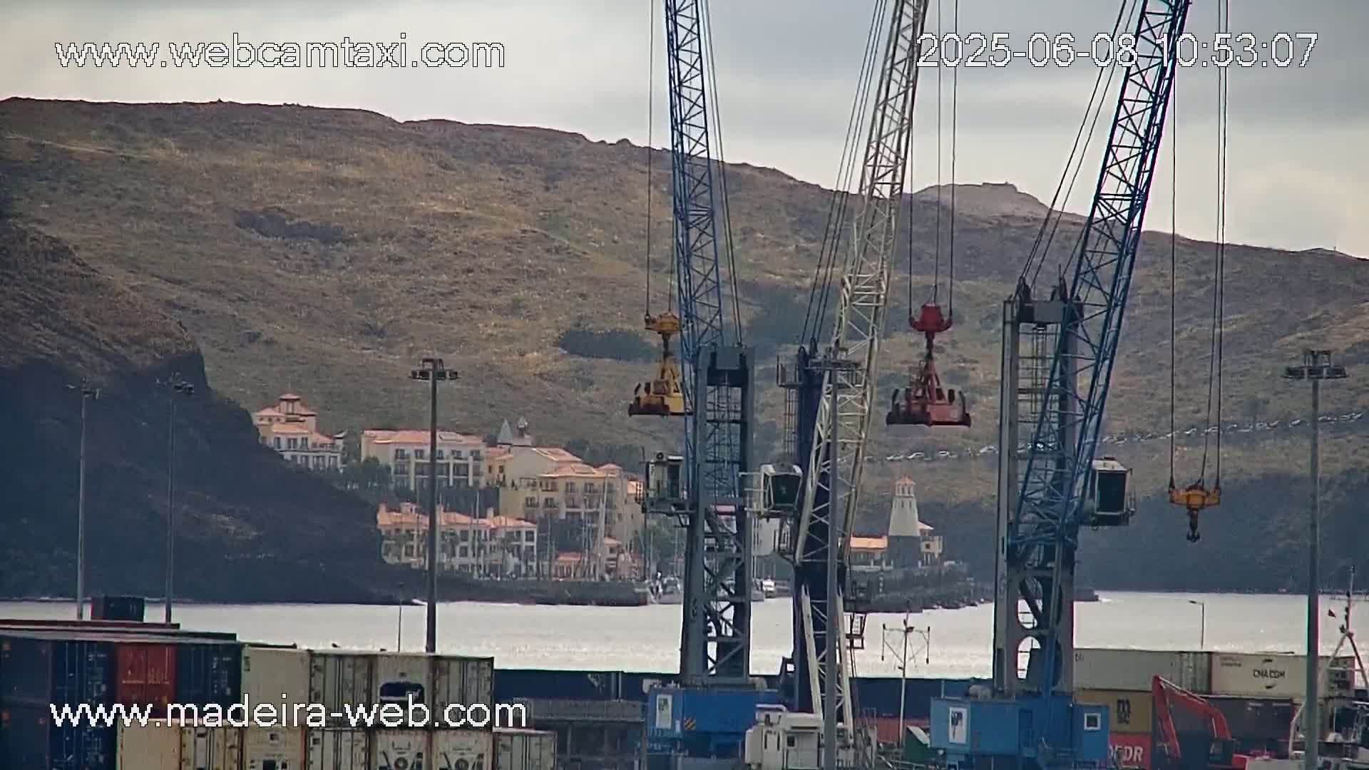 Several large cranes are positioned in a harbor, overlooking a town nestled against a hillside under an overcast sky.