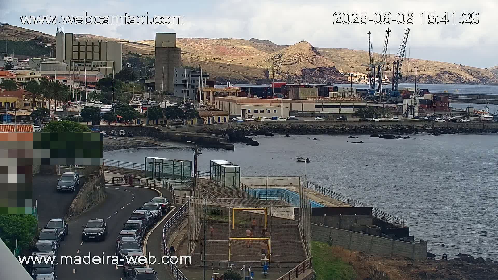 A coastal town with a harbor, featuring industrial buildings, cars on a road, a small beach, and people playing soccer on a nearby field, under partly cloudy skies.