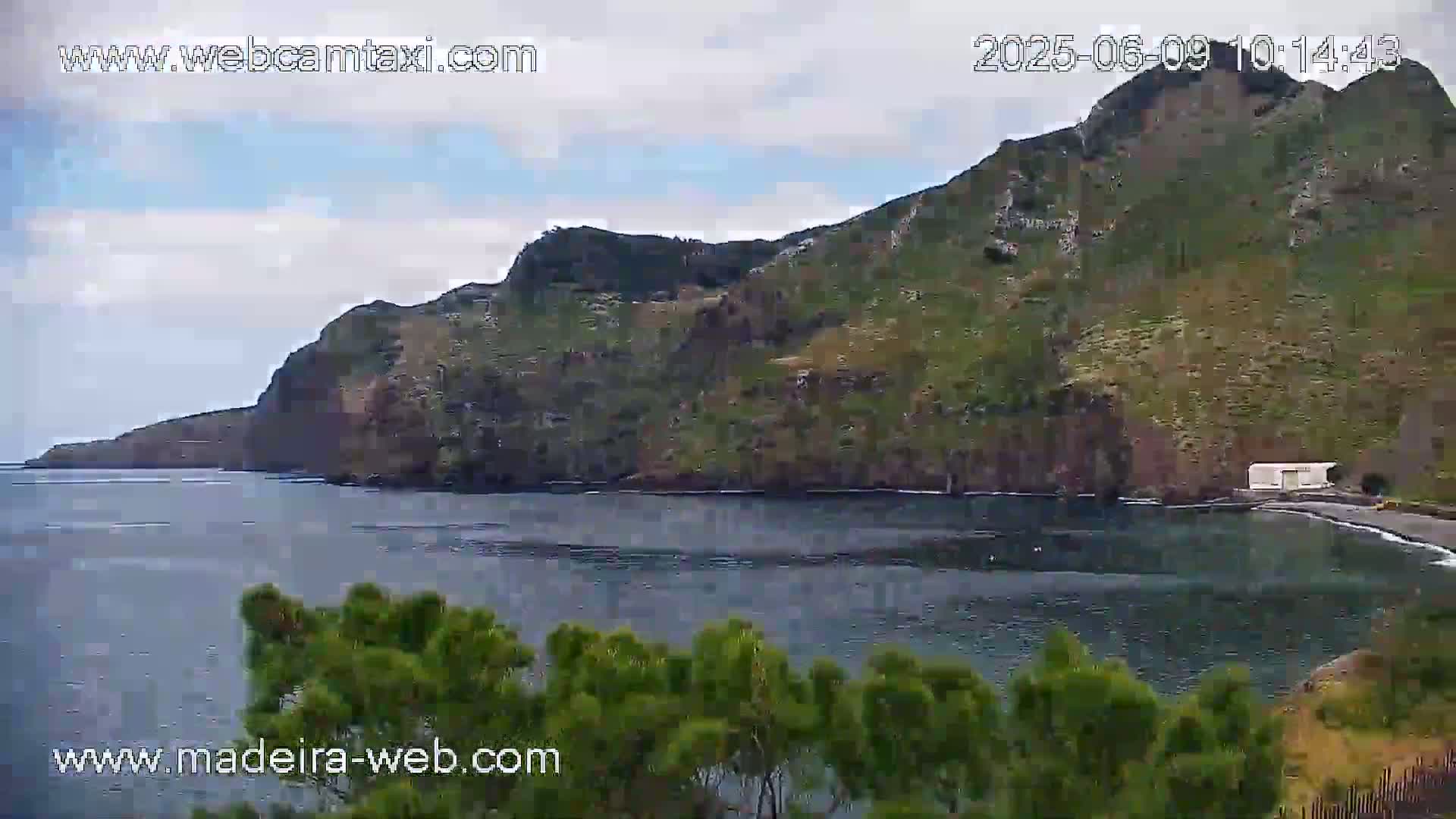 A rocky, green-covered hillside slopes down to a calm, dark-blue bay under a mostly cloudy sky.