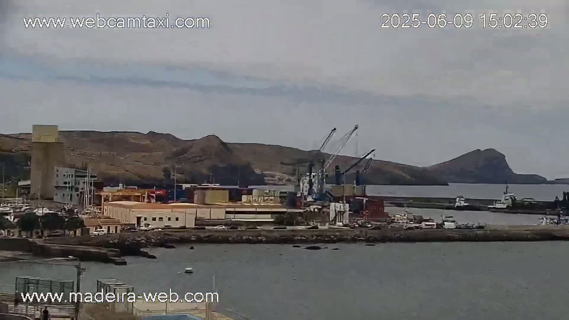A harbor scene under an overcast sky shows cargo containers, cranes, and several boats near a rocky shoreline and hills.