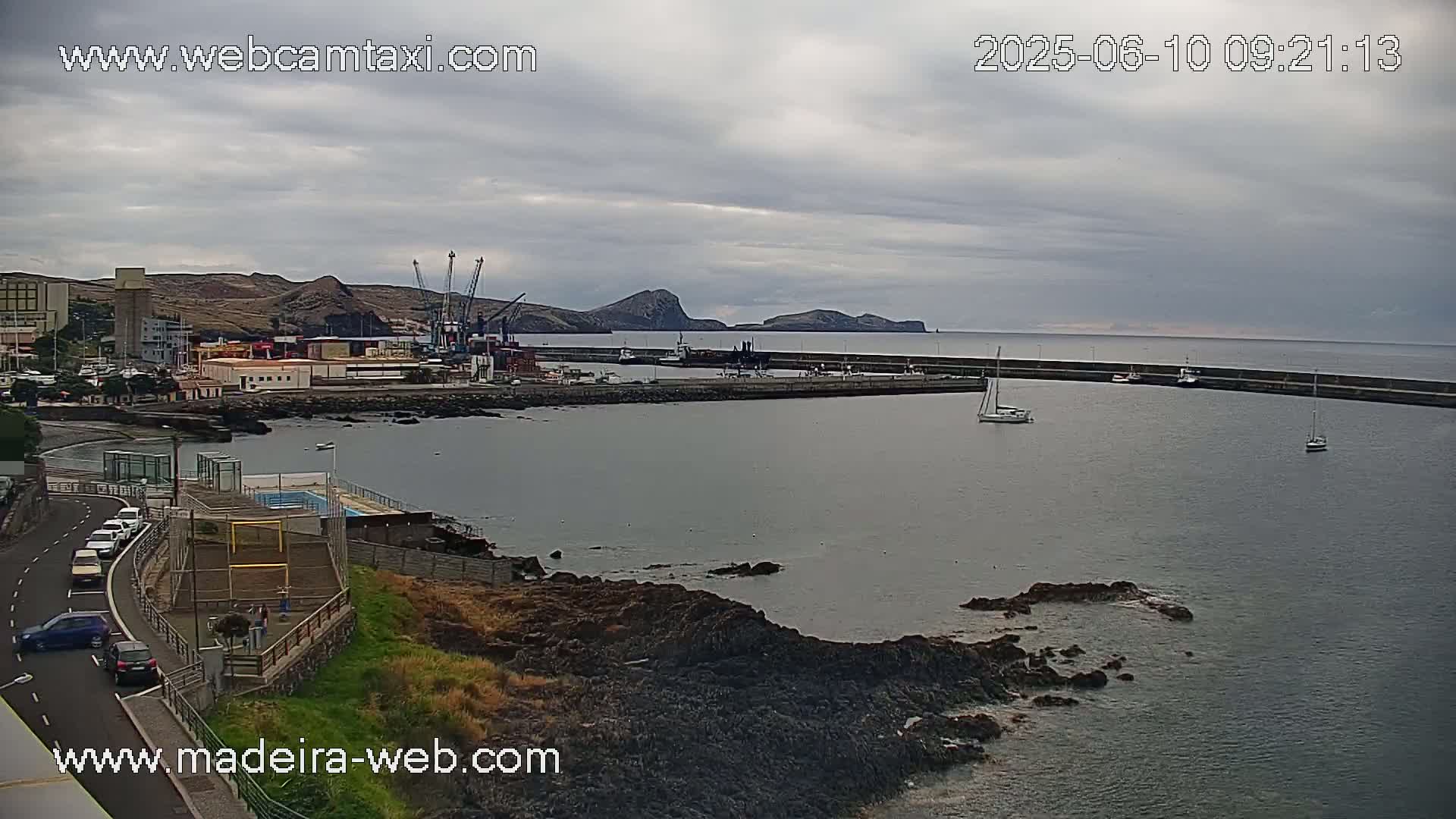A harbor with a few boats is visible under an overcast sky, with a rocky shoreline and buildings in the background.