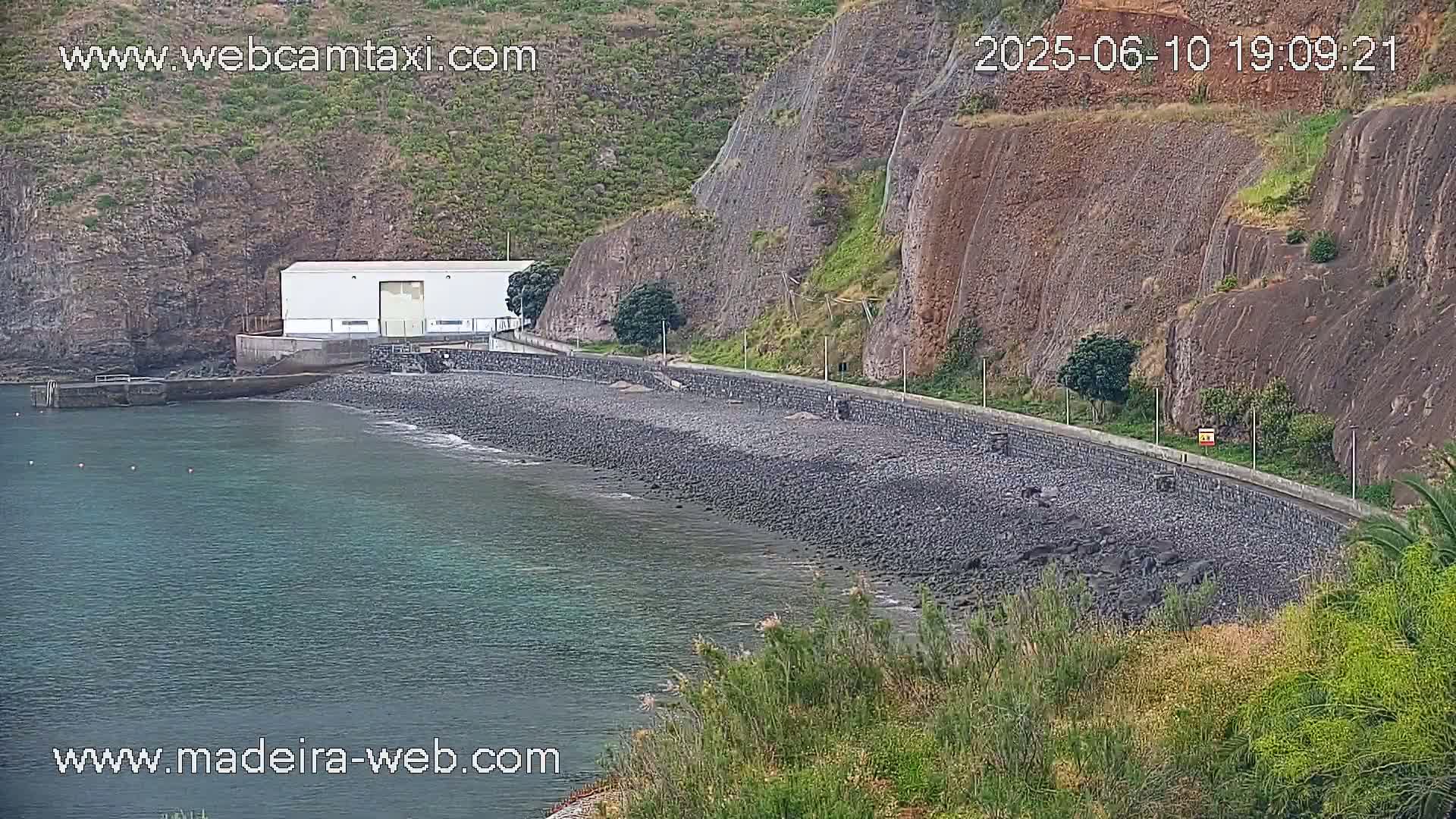 A white building sits near a rocky, dark-colored beach next to a steep, green cliff overlooking calm, dark-green water.