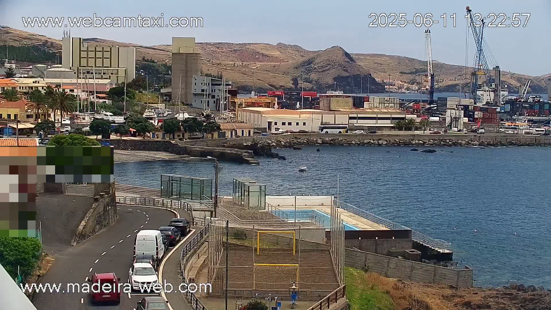 A coastal town scene on a sunny day shows a harbor with boats, cargo containers, and industrial buildings, along with a nearby road, small swimming pool, and soccer goals.