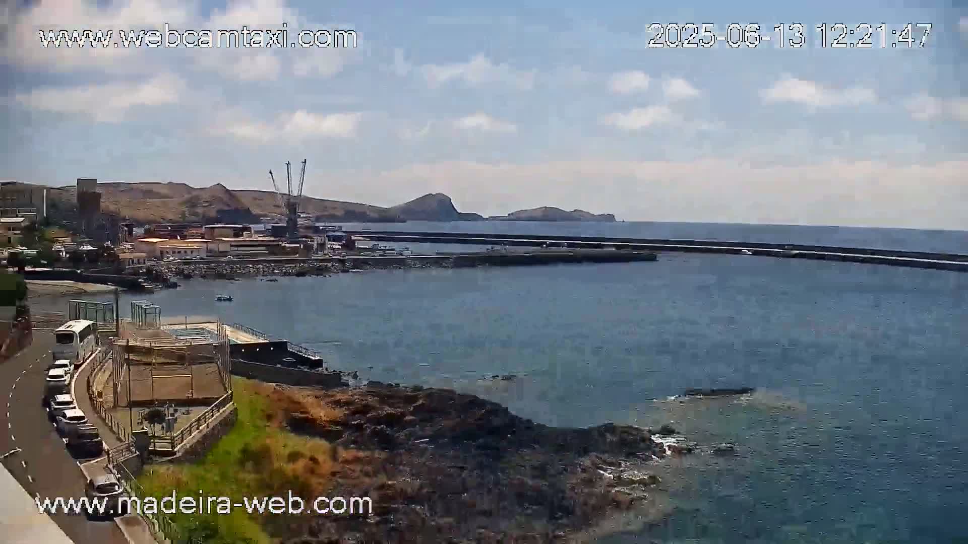 A partly sunny day shows a harbor town with a calm sea and a breakwater, cars parked along a road, and a bus on a nearby street.