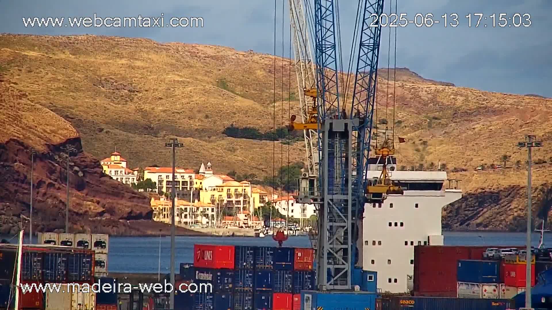 A large crane stands near a cargo ship and stacks of shipping containers in a harbor, with a hillside town and dry, tan-colored mountains in the background under a mostly clear sky.