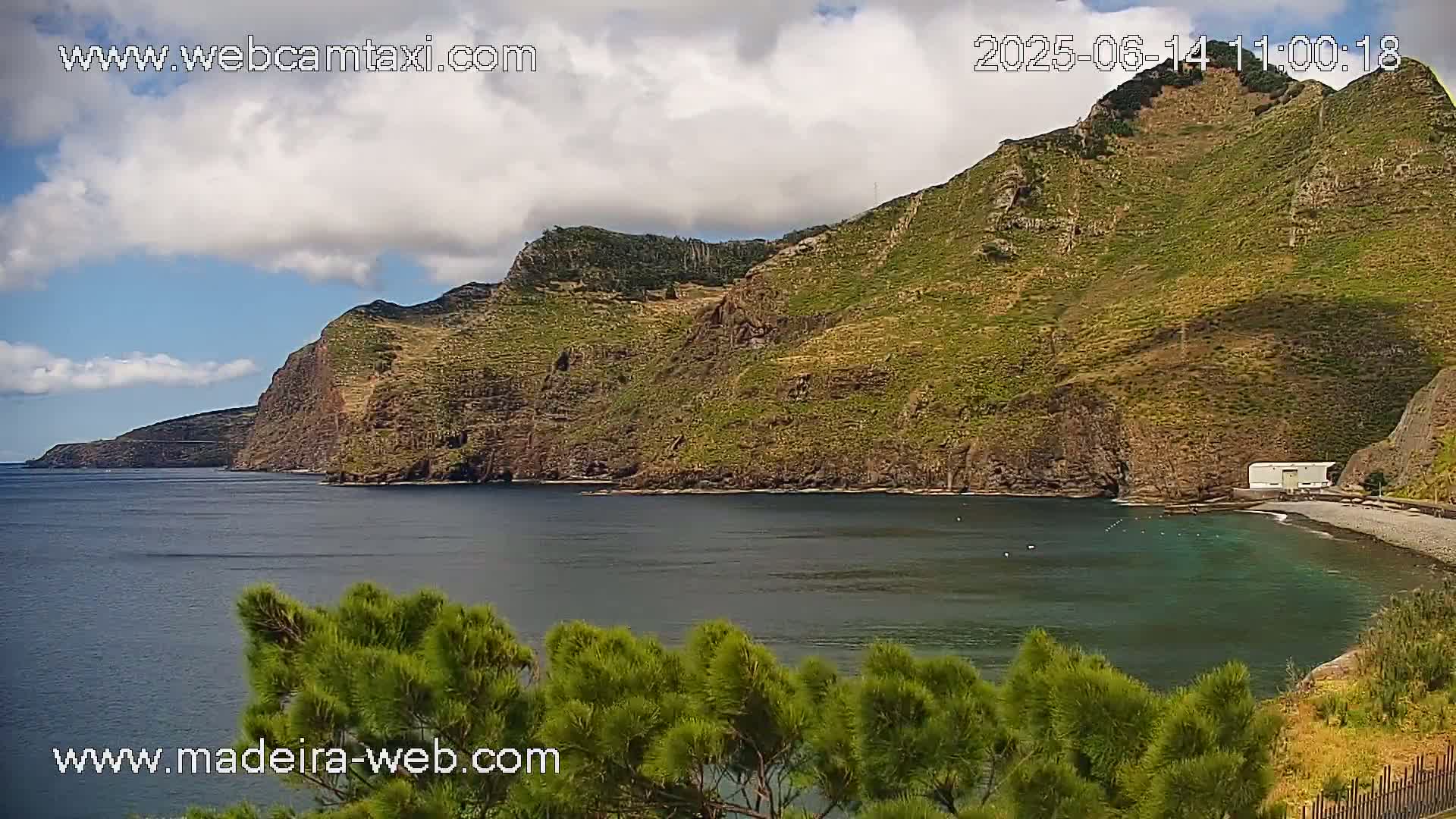 A partly cloudy day reveals a rocky, green-covered cliffside meeting a calm, dark-blue ocean, with a small beach visible in the foreground.