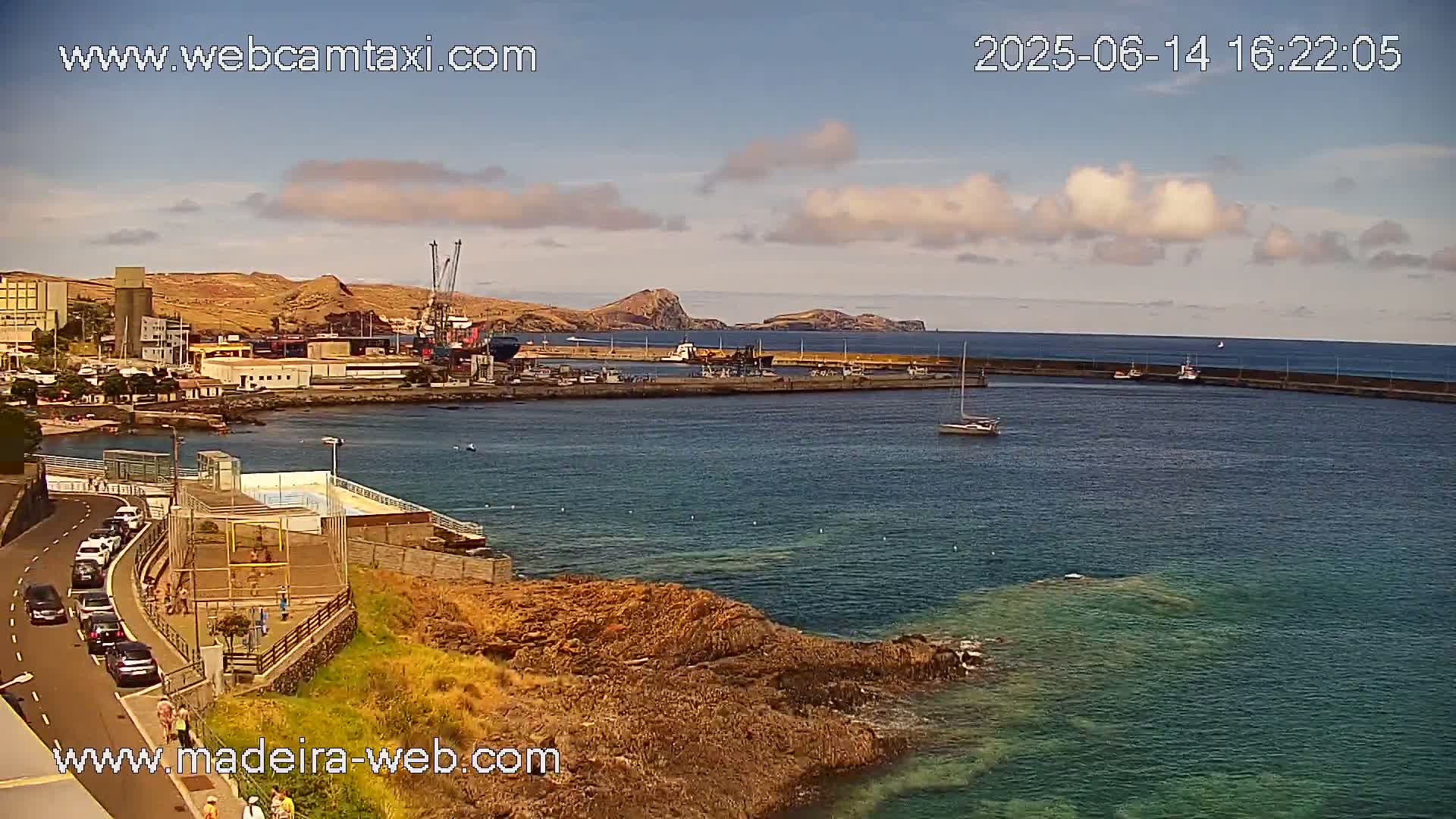 A partly sunny day shows a harbor with boats, a small town, and a rocky coastline.