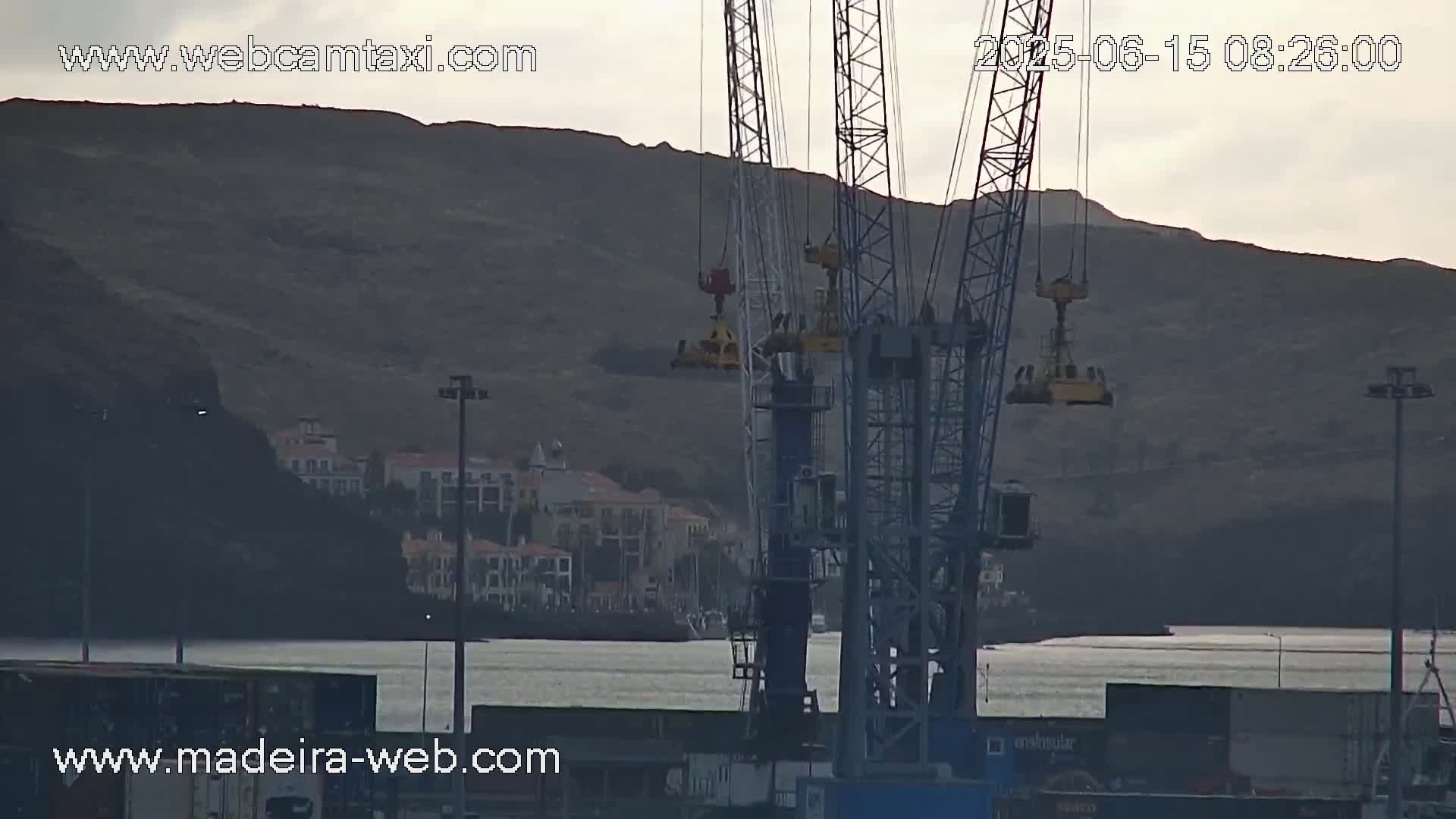 Two large cranes are positioned in a harbor, overlooking a calm body of water and a town nestled against a hill under an overcast sky.