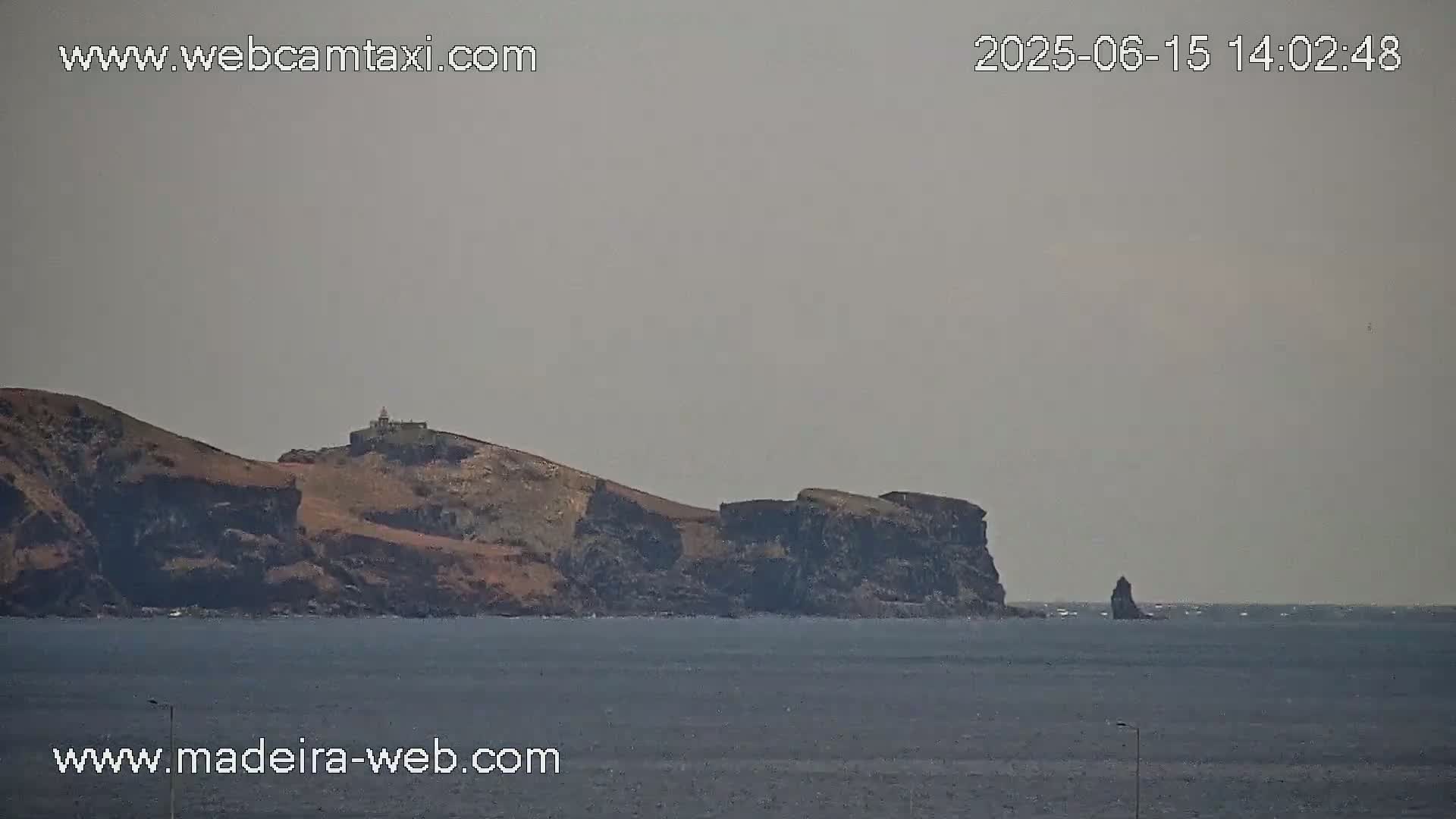 A hazy view shows a rocky island with a small building on top, extending into a calm ocean with a solitary rock formation further out.