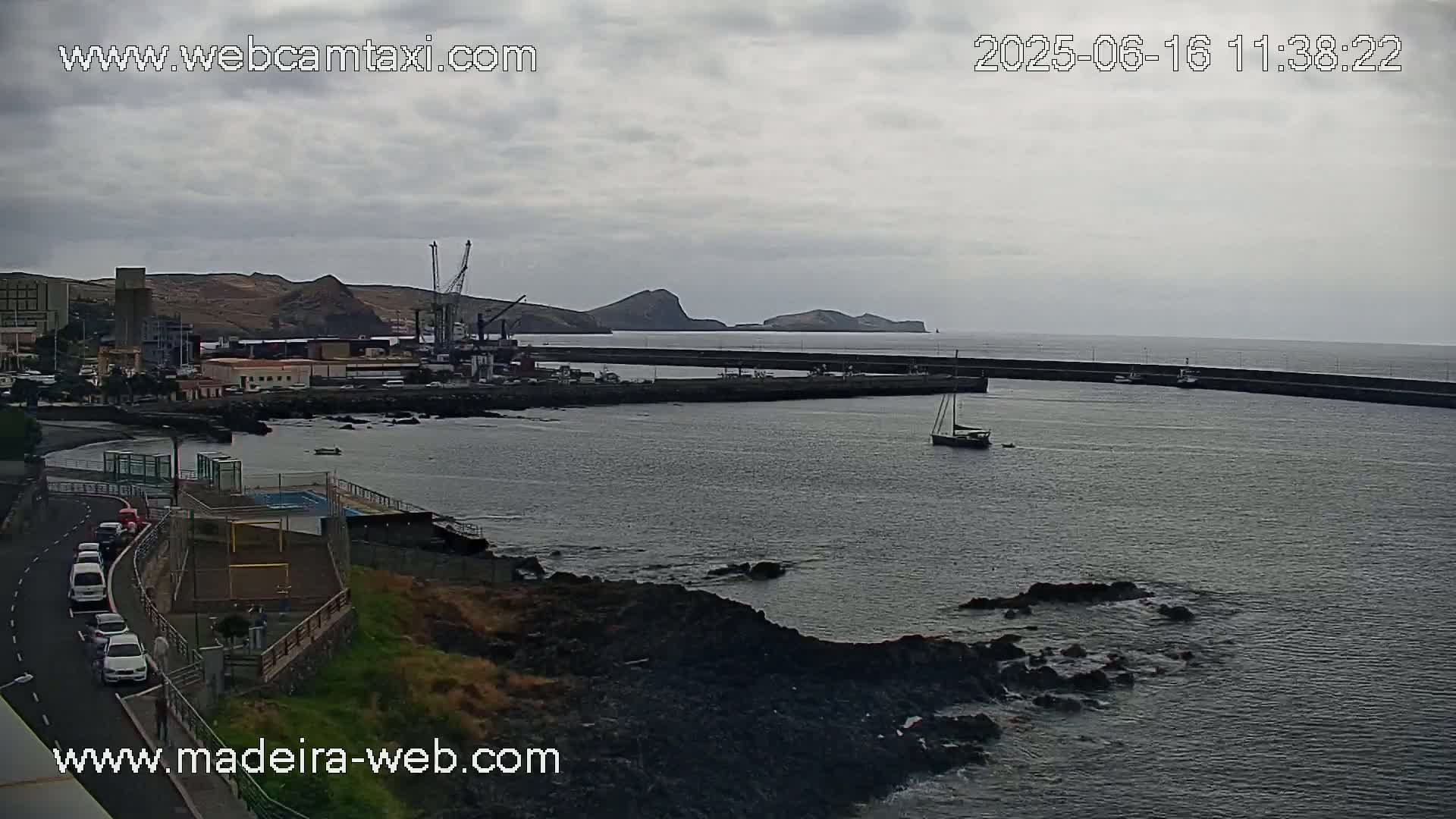 A harbor scene under an overcast sky shows a small sailboat in calm water near a breakwater, with a town, industrial buildings, and distant hills visible in the background.