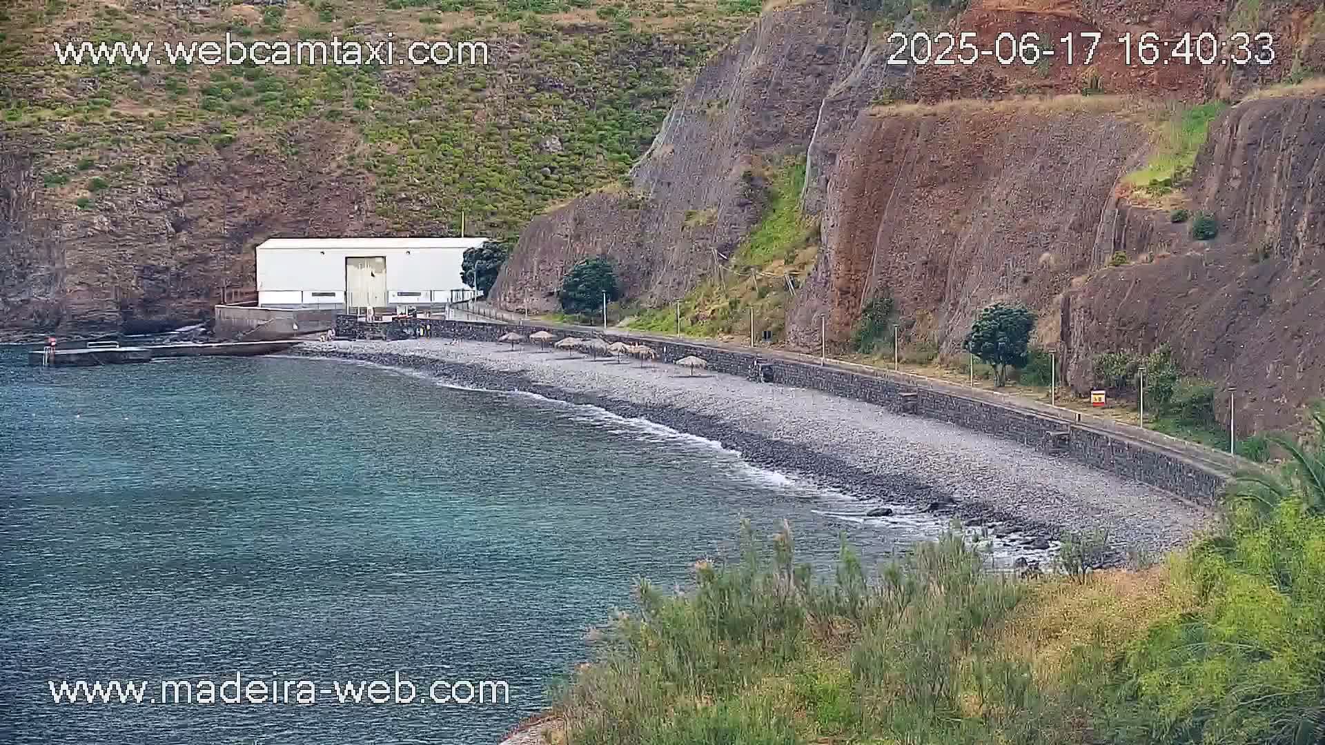 A rocky beach with calm ocean water laps a shore next to a white building against a backdrop of dark cliffs.