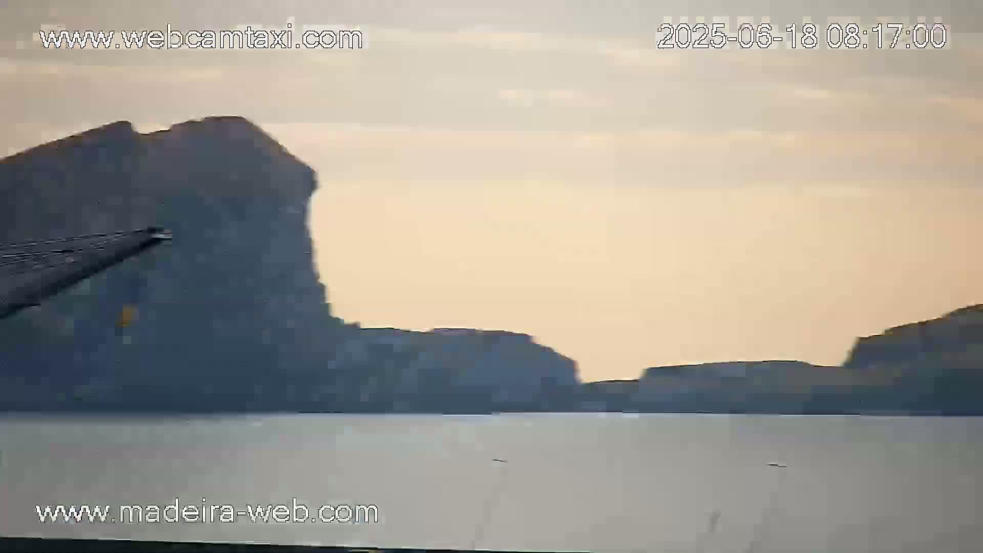 A calm body of water stretches out before a range of dark, rocky cliffs under a pale, hazy sky.