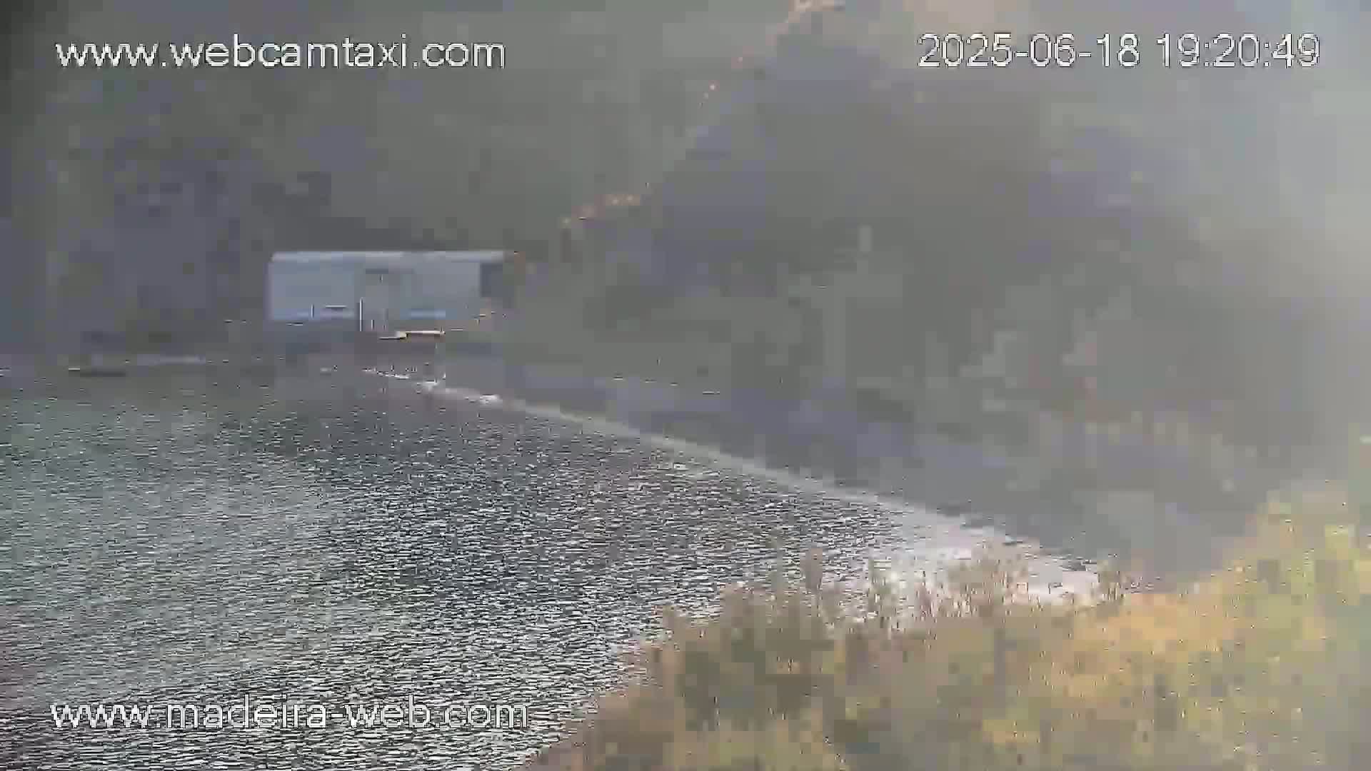 A hazy view shows a body of water next to a shoreline with sparse vegetation, and a small building in the background.
