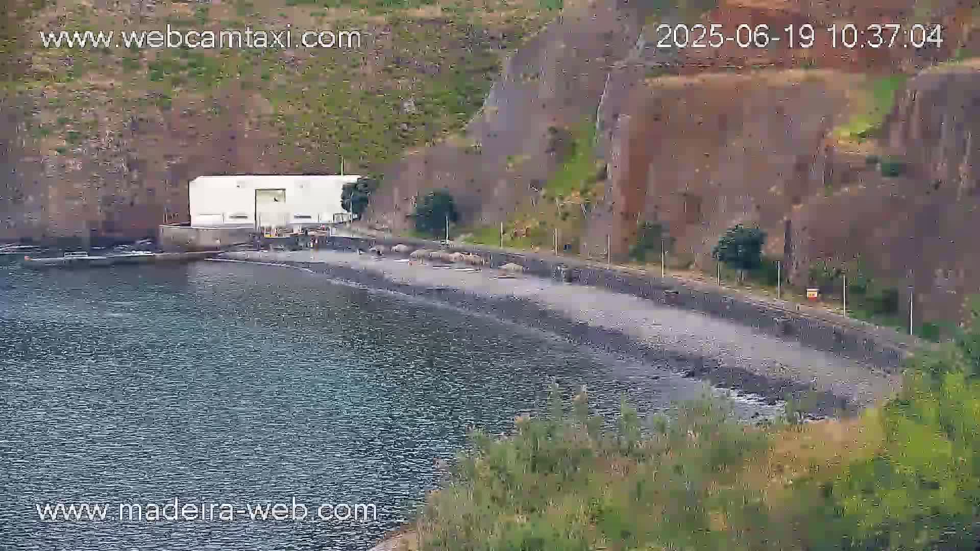 A dark-colored, rocky beach curves around a calm body of water next to a white building situated against a steep, rocky cliffside.