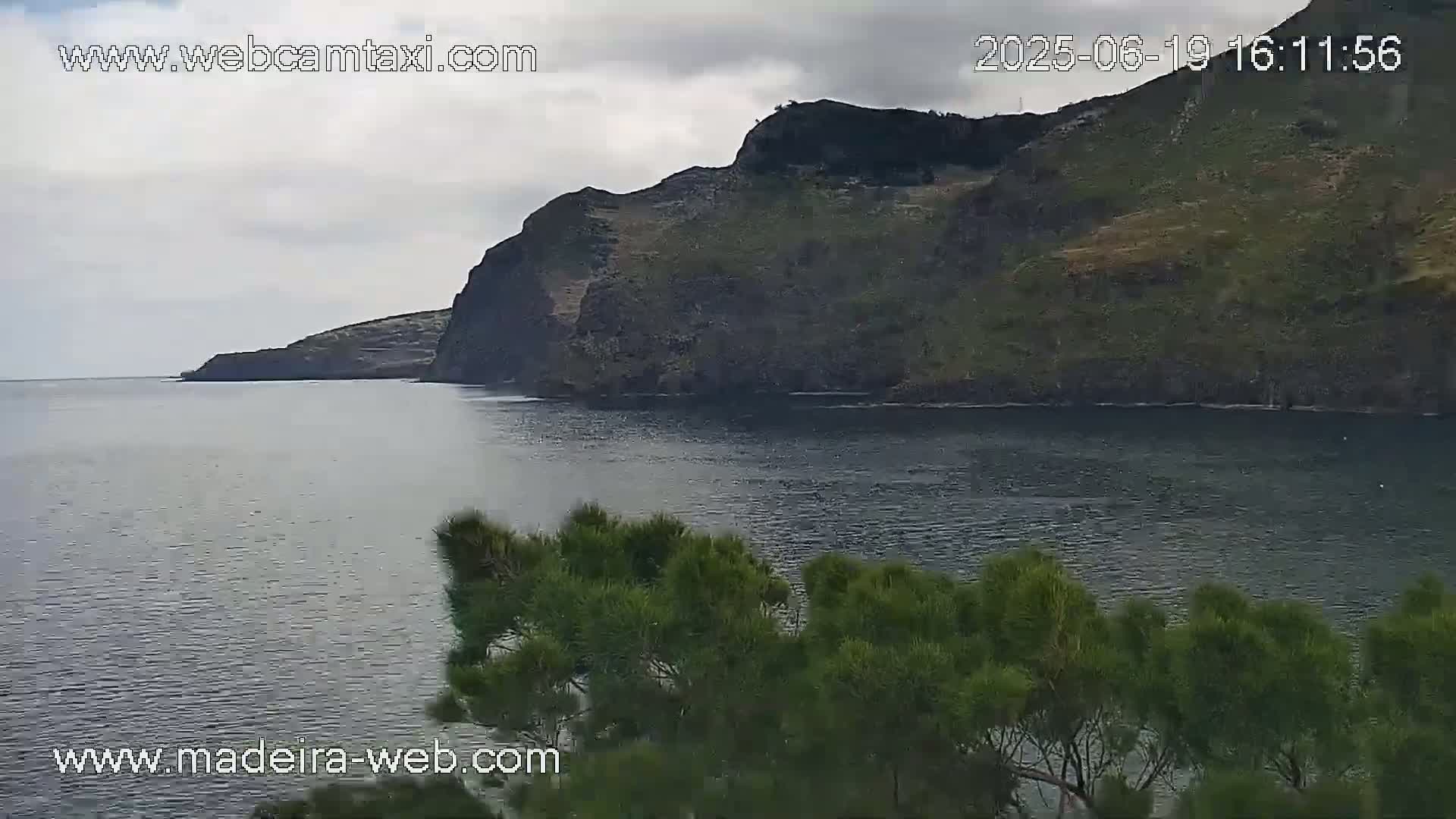 A dark, calm body of water stretches out before a rocky, green-covered cliff under a cloudy sky.