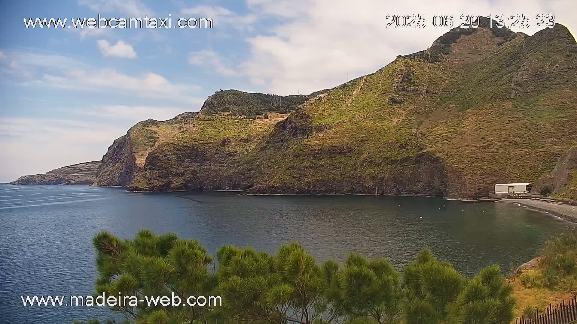 A partly cloudy day shows a tranquil cove nestled between steep, green hillsides, a small beach at the base, and dark blue water.