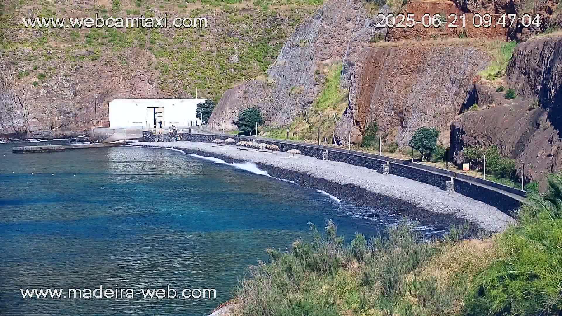 A rocky, dark-colored beach curves along a calm, blue ocean under a sunny sky, with a white building and a low wall bordering the shore.
