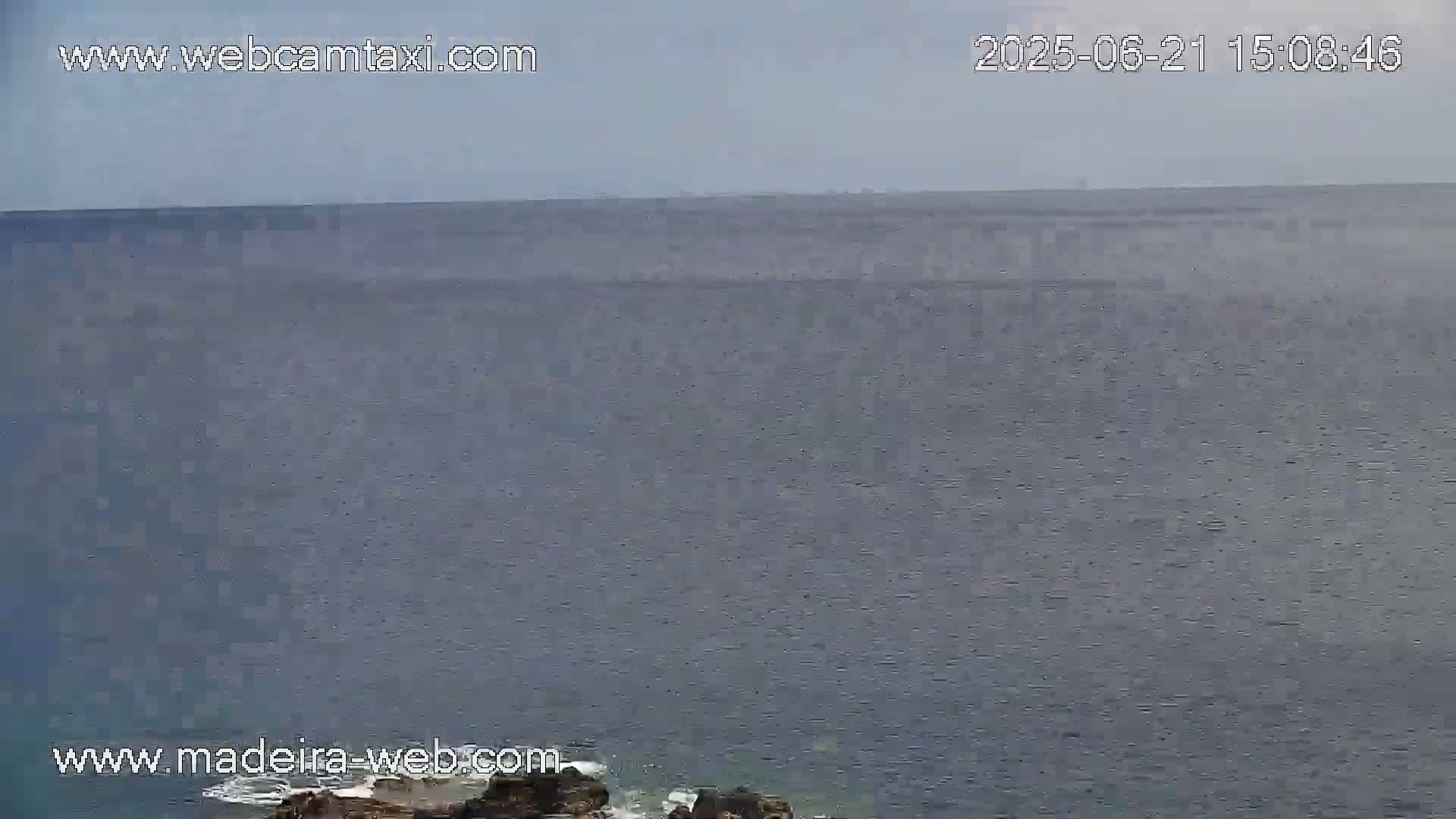 A calm, gray ocean is seen from a high vantage point, with dark rocks visible at the bottom edge of the frame.