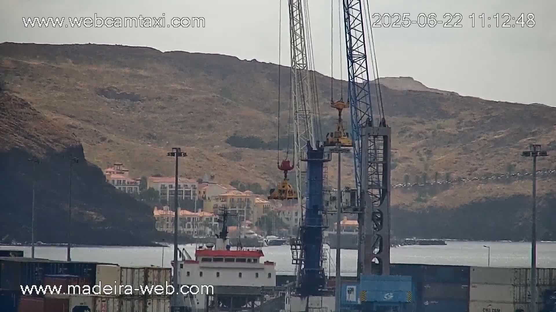 Two large cranes are operating near a harbor, with buildings and a hill visible in the background under hazy skies.
