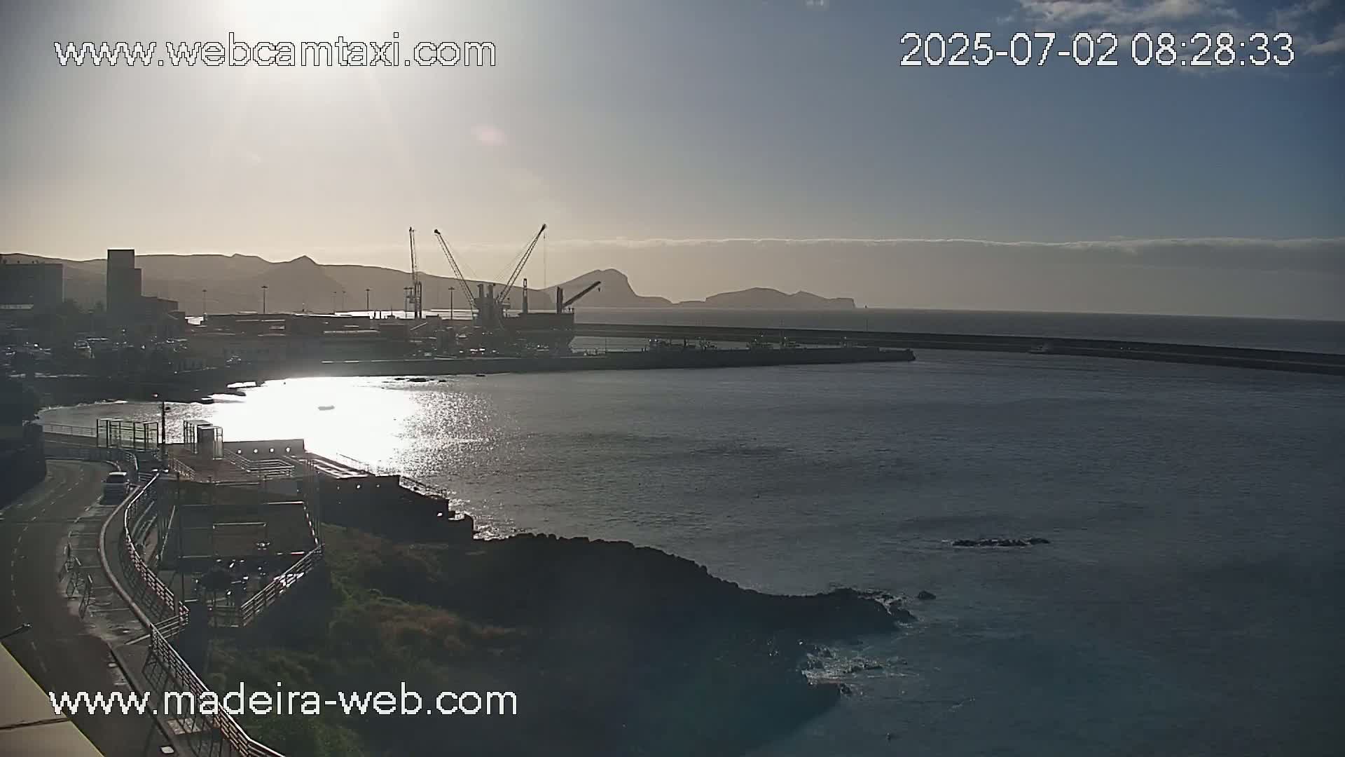 A harbor with a breakwater, cargo ships, and cranes is seen at sunrise under mostly clear skies.