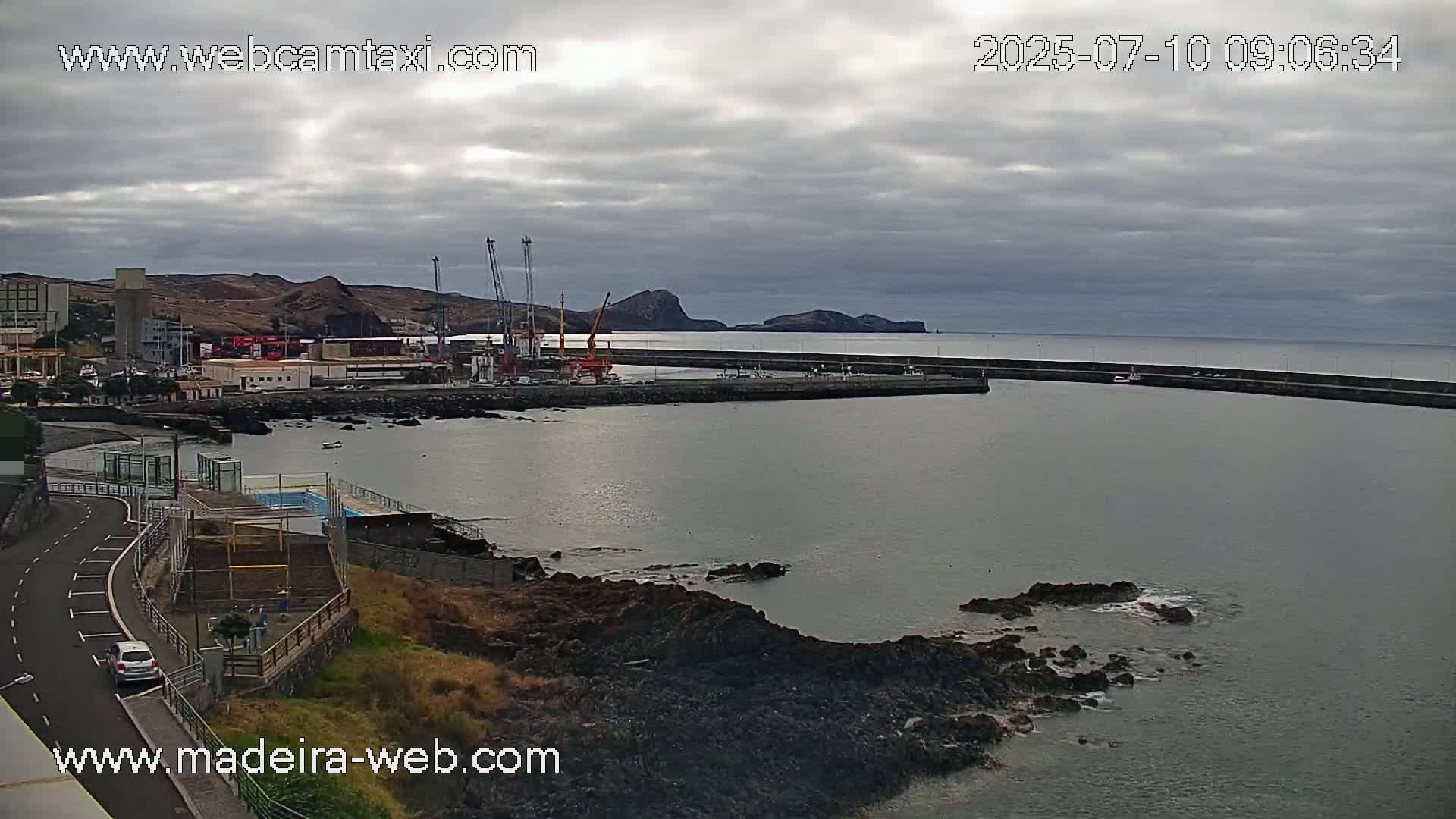 A harbor with overcast skies shows a small town nestled beside a rocky coast with calm water and several cranes at a dock.