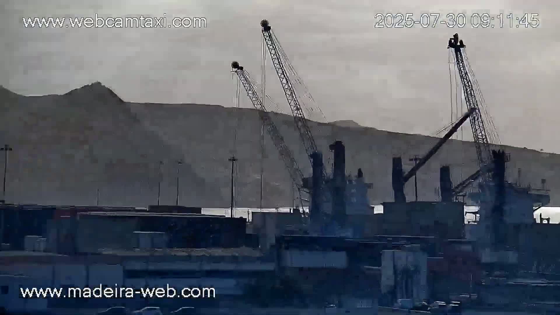 A hazy, overcast day reveals several large cranes and industrial structures at a port, with a hill visible in the background.