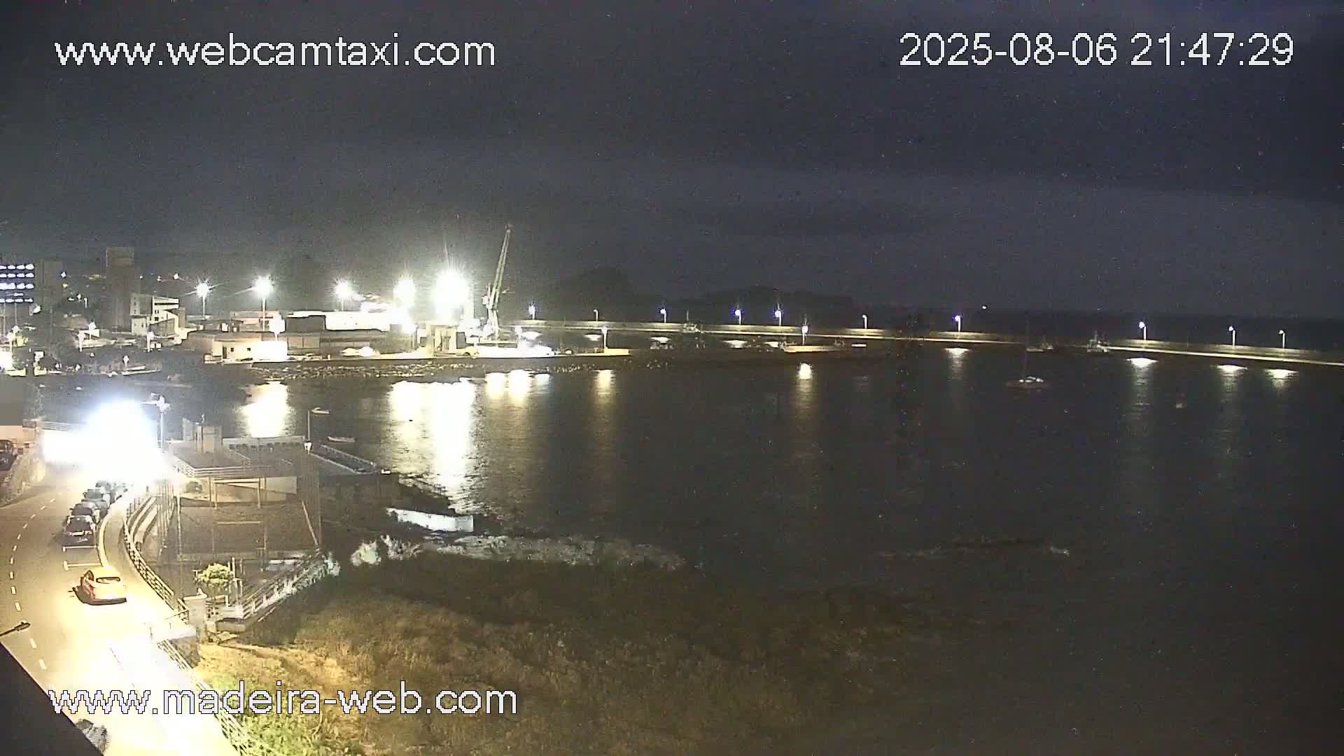 A nighttime view of a harbor with calm water, illuminated by lights along a seawall, showing a few boats and industrial structures along the shore.