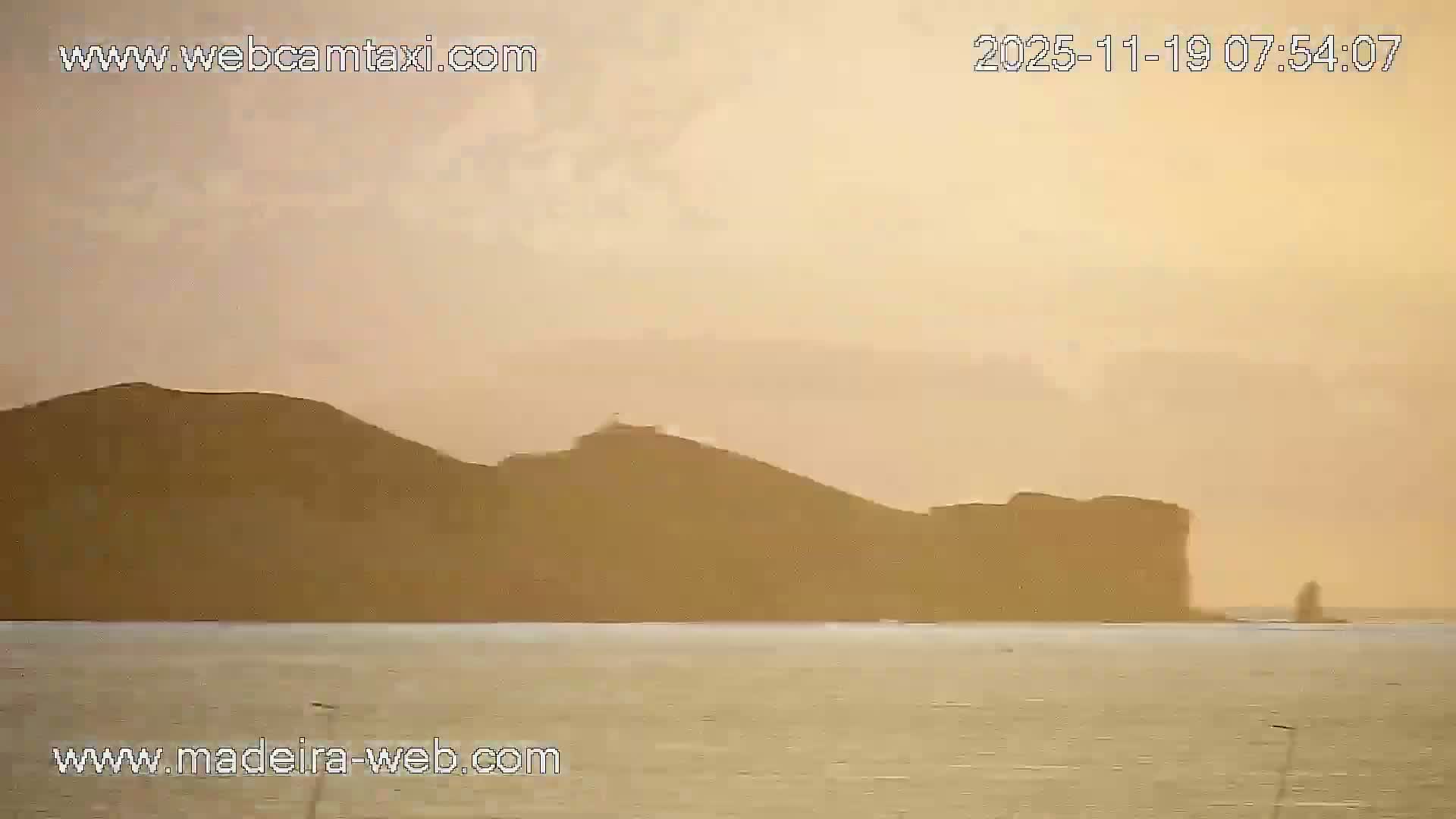 A nighttime view of a harbor with calm water, illuminated by lights along a seawall, showing a few boats and industrial structures along the shore.