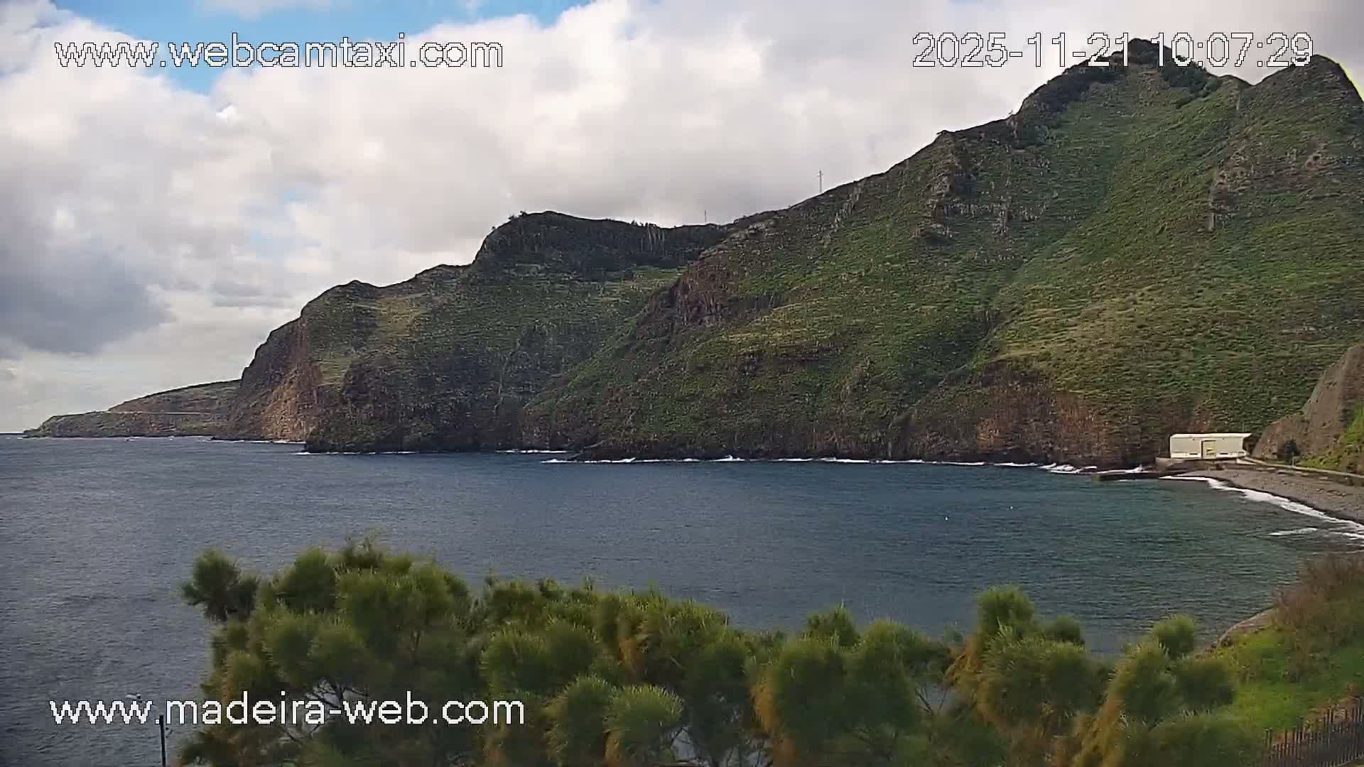 The image shows a dramatic coastal landscape with steep, verdant cliffs descending into a dark blue sea, a pebbly beach featuring a white building at its edge, and a partly cloudy sky overhead.