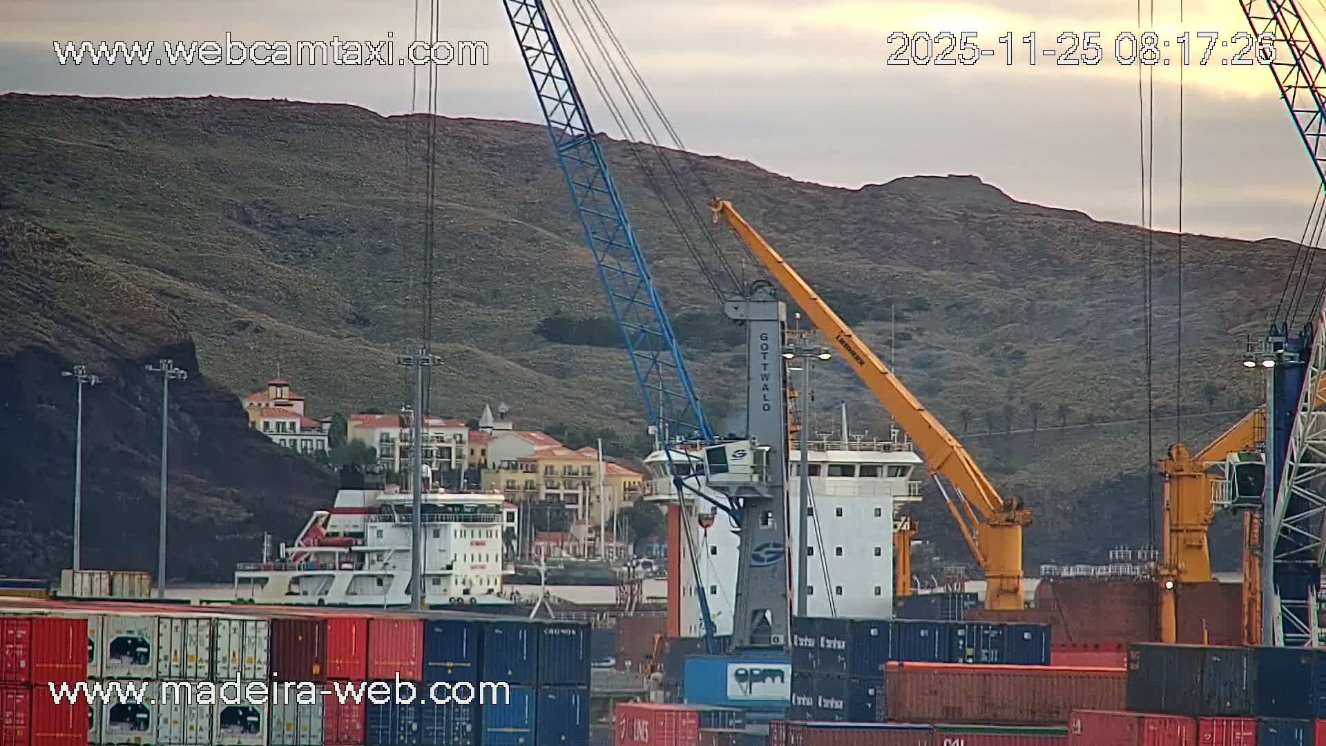 Under a cloudy morning sky, a bustling port scene unfolds with stacks of colorful shipping containers, several large cranes, and ships docked against steep, green hills dotted with buildings.