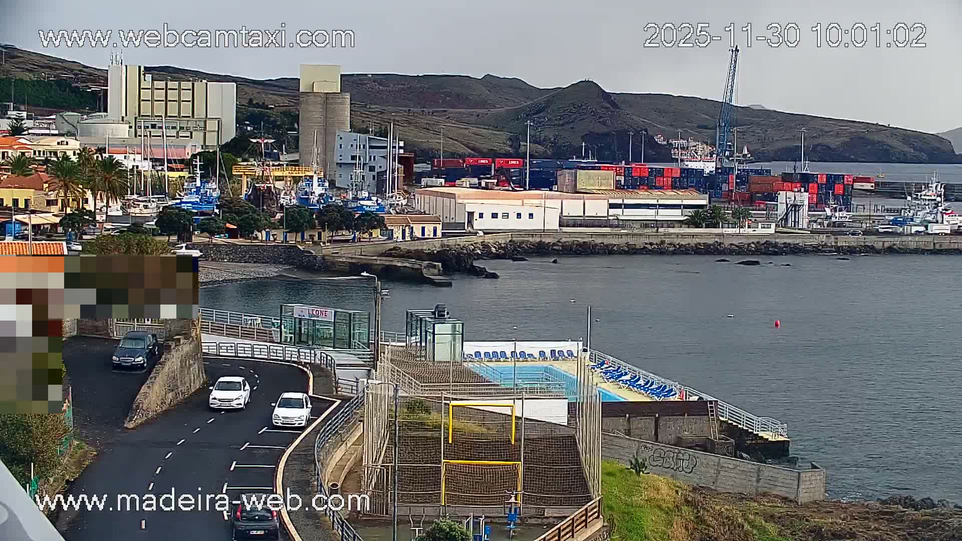 The image shows an overcast day over a bustling coastal town featuring a harbor with various boats and cargo ships, industrial buildings, a winding road with cars, and recreational facilities including a swimming pool and sports field, all set against a backdrop of hills.