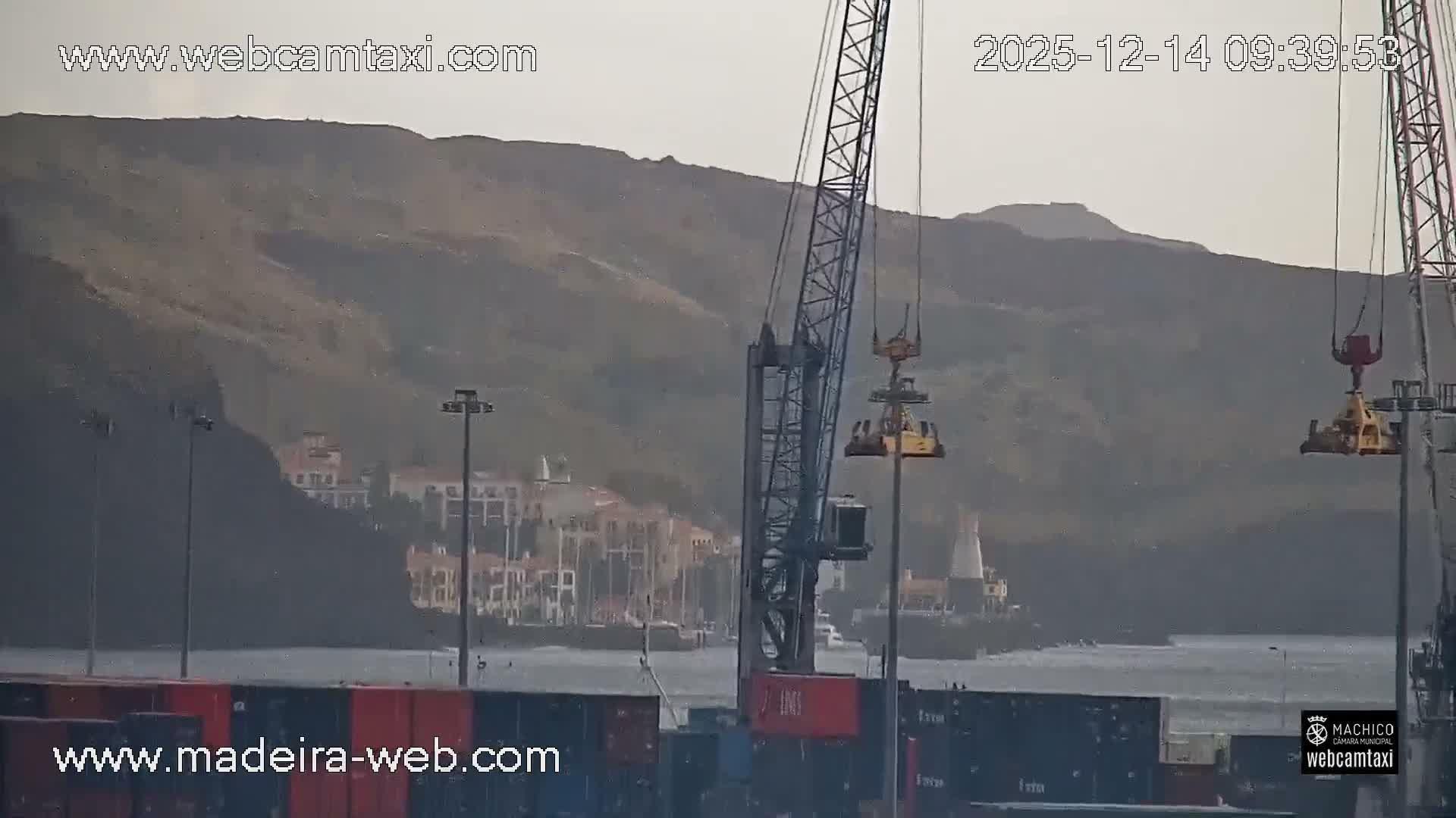 The image shows an overcast day over a bustling coastal town featuring a harbor with various boats and cargo ships, industrial buildings, a winding road with cars, and recreational facilities including a swimming pool and sports field, all set against a backdrop of hills.