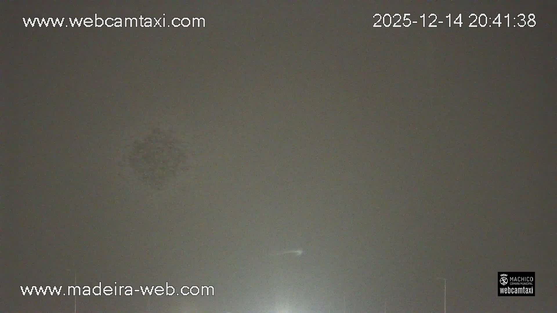 The image shows an overcast day over a bustling coastal town featuring a harbor with various boats and cargo ships, industrial buildings, a winding road with cars, and recreational facilities including a swimming pool and sports field, all set against a backdrop of hills.