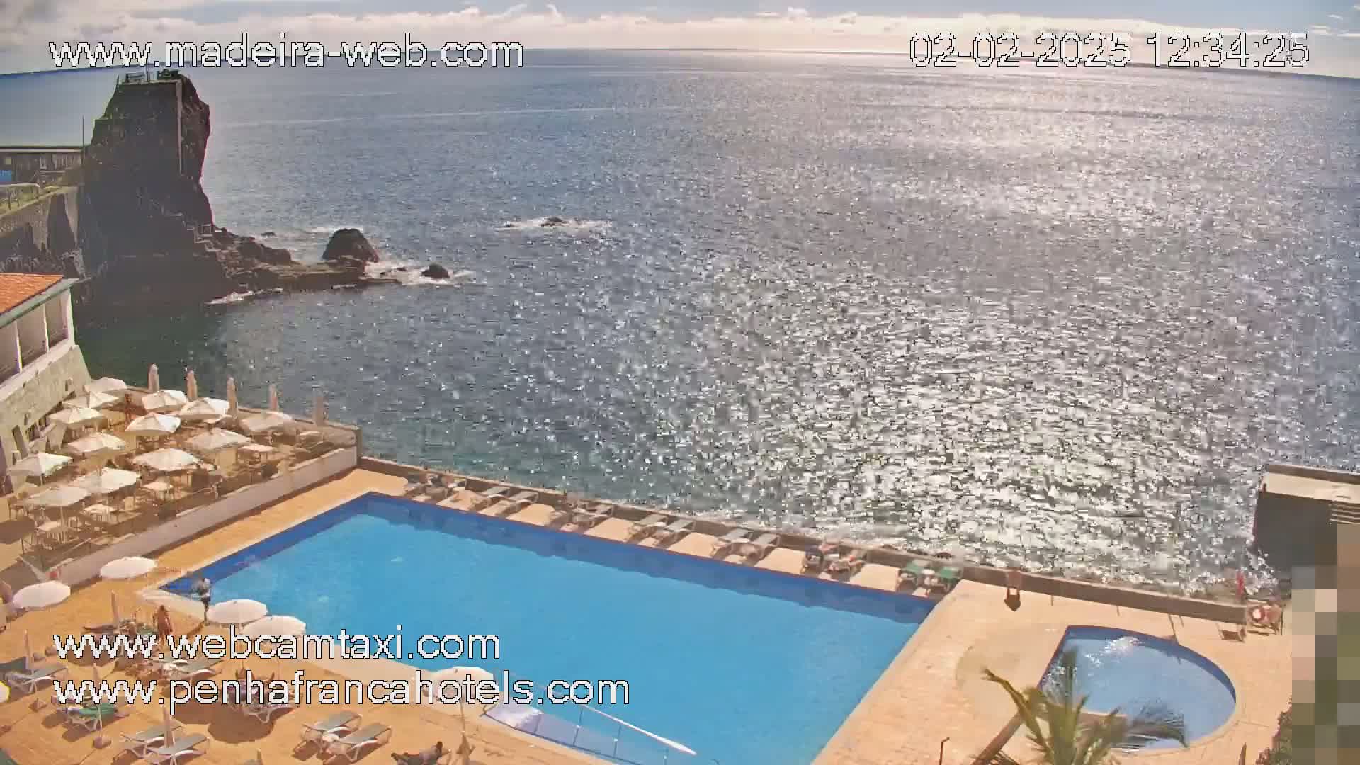 A hotel pool area with lounge chairs and umbrellas overlooks a sparkling ocean next to a rocky outcrop under a partly cloudy sky.