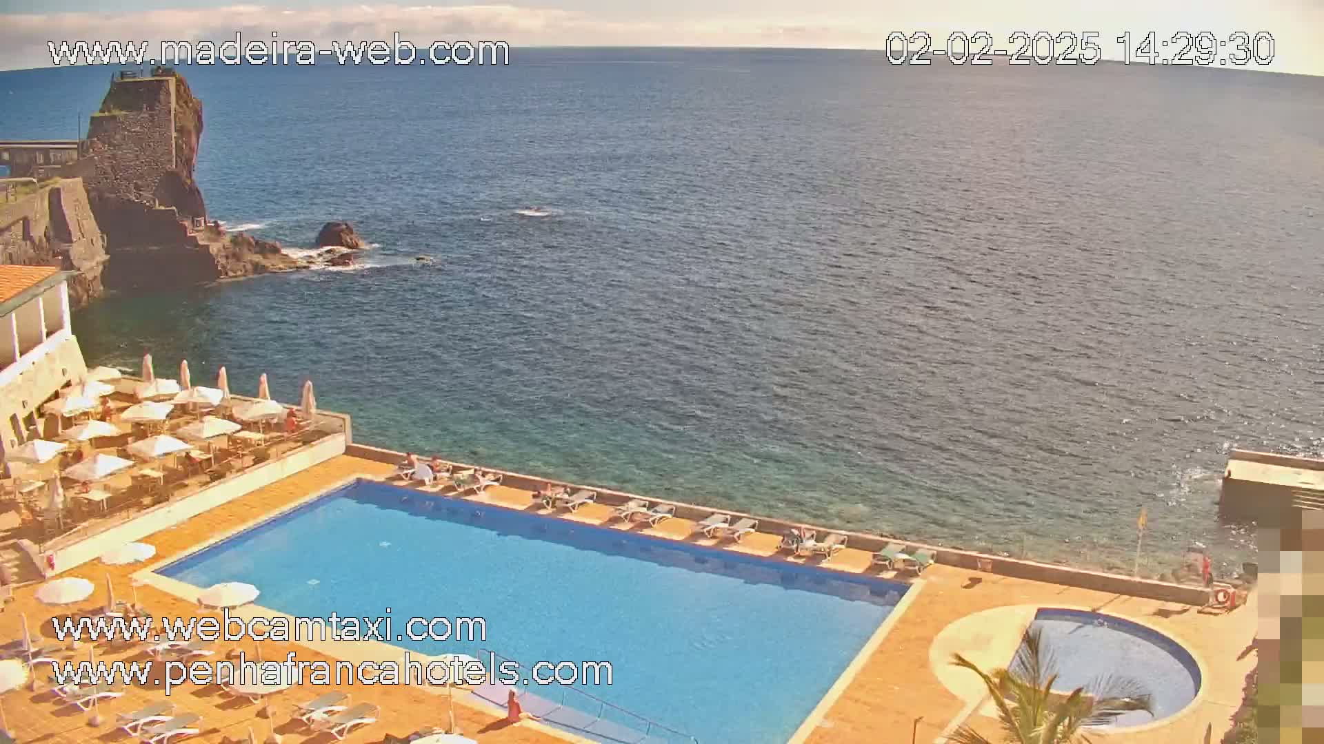 A resort pool and patio area overlooks a calm ocean on a sunny day.