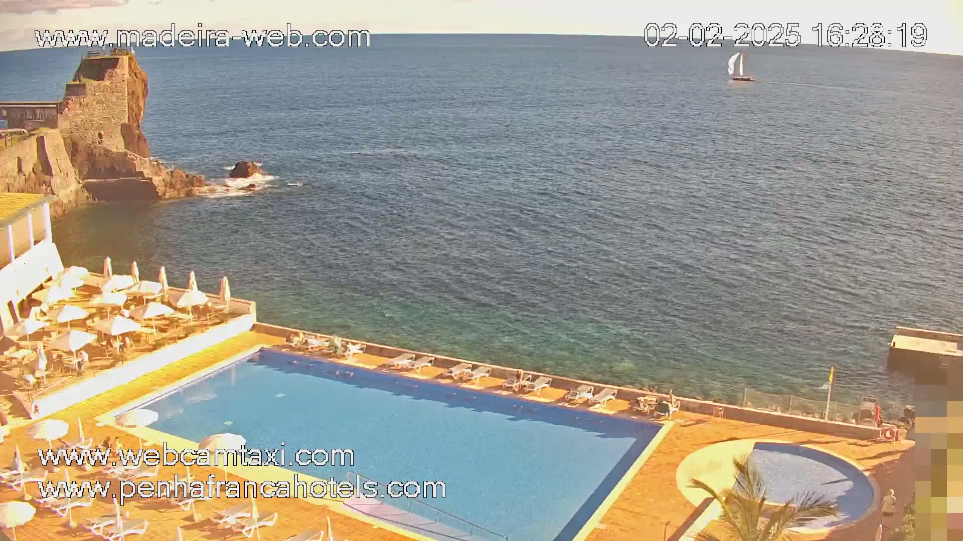 A hotel pool and patio area overlook a calm ocean with a sailboat in the distance under a sunny sky.