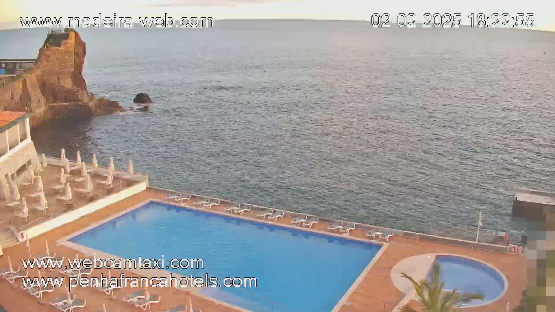 A resort pool area with lounge chairs and umbrellas overlooks a calm ocean with a rocky outcrop in the distance.