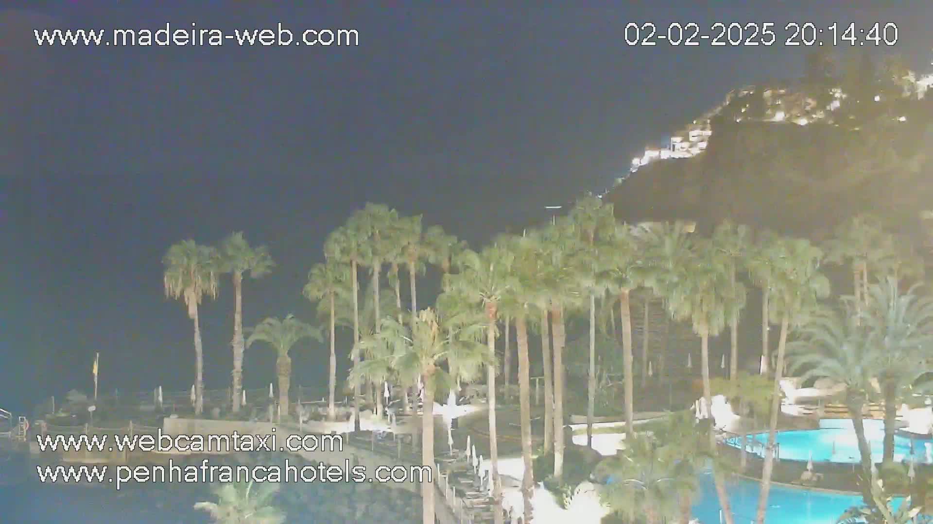 A nighttime view of a resort area with palm trees surrounding a swimming pool, illuminated buildings on a hillside in the background under a dark sky.