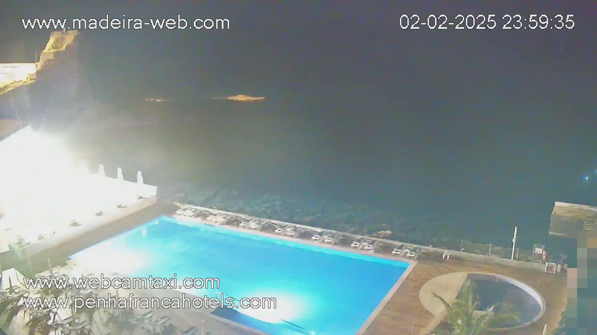 An outdoor nighttime view shows a brightly lit swimming pool next to a dark ocean under a clear, dark sky.