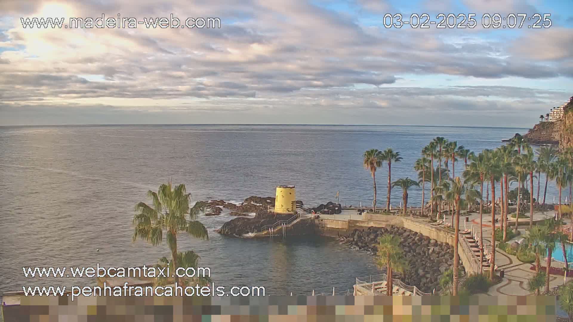 A partly cloudy sky overlooks a calm ocean, a rocky shoreline with palm trees, and a small yellow tower near a swimming pool.