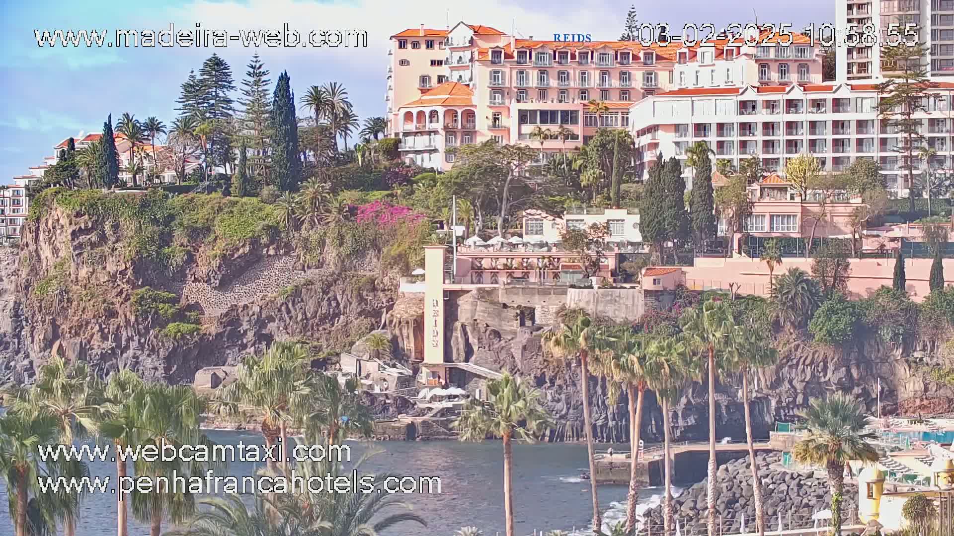 A coastal scene on a sunny day shows several hotels situated on a cliff overlooking a dark body of water with palm trees and lush vegetation.